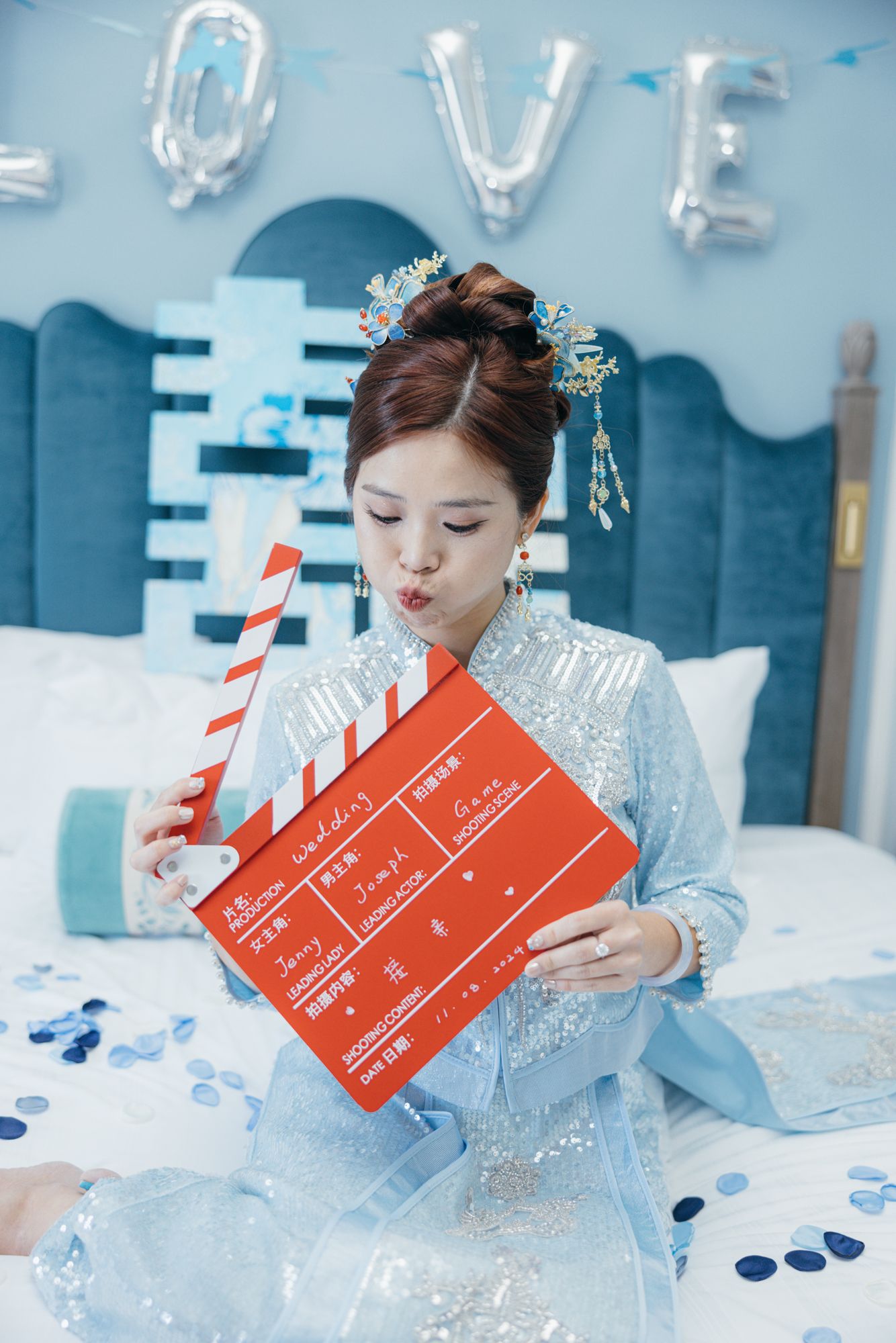Bride in sequined blue qipao holds red wedding clapperboard on decorated bed with LOVE balloons.