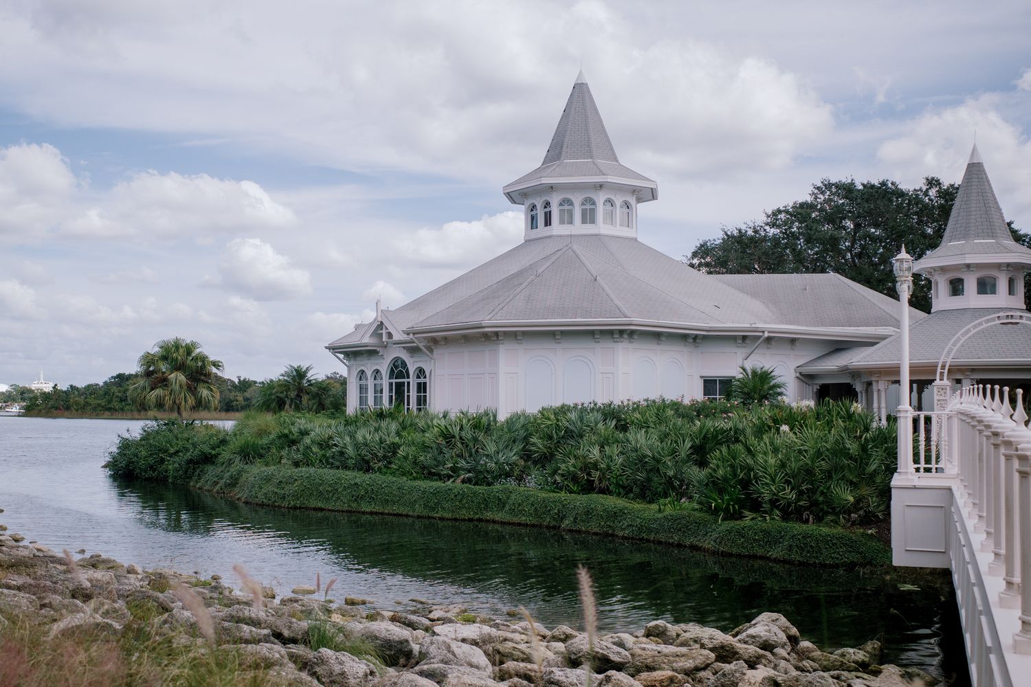 White Victorian-style waterfront chapel with gray roof surrounded by lush greenery and rocky shoreline under cloudy skies.