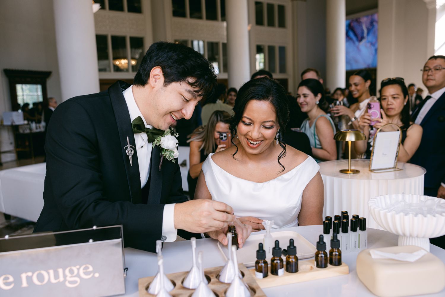 Smiling bride and groom mix custom fragrance oils at a wedding reception table while guests watch from nearby tables.