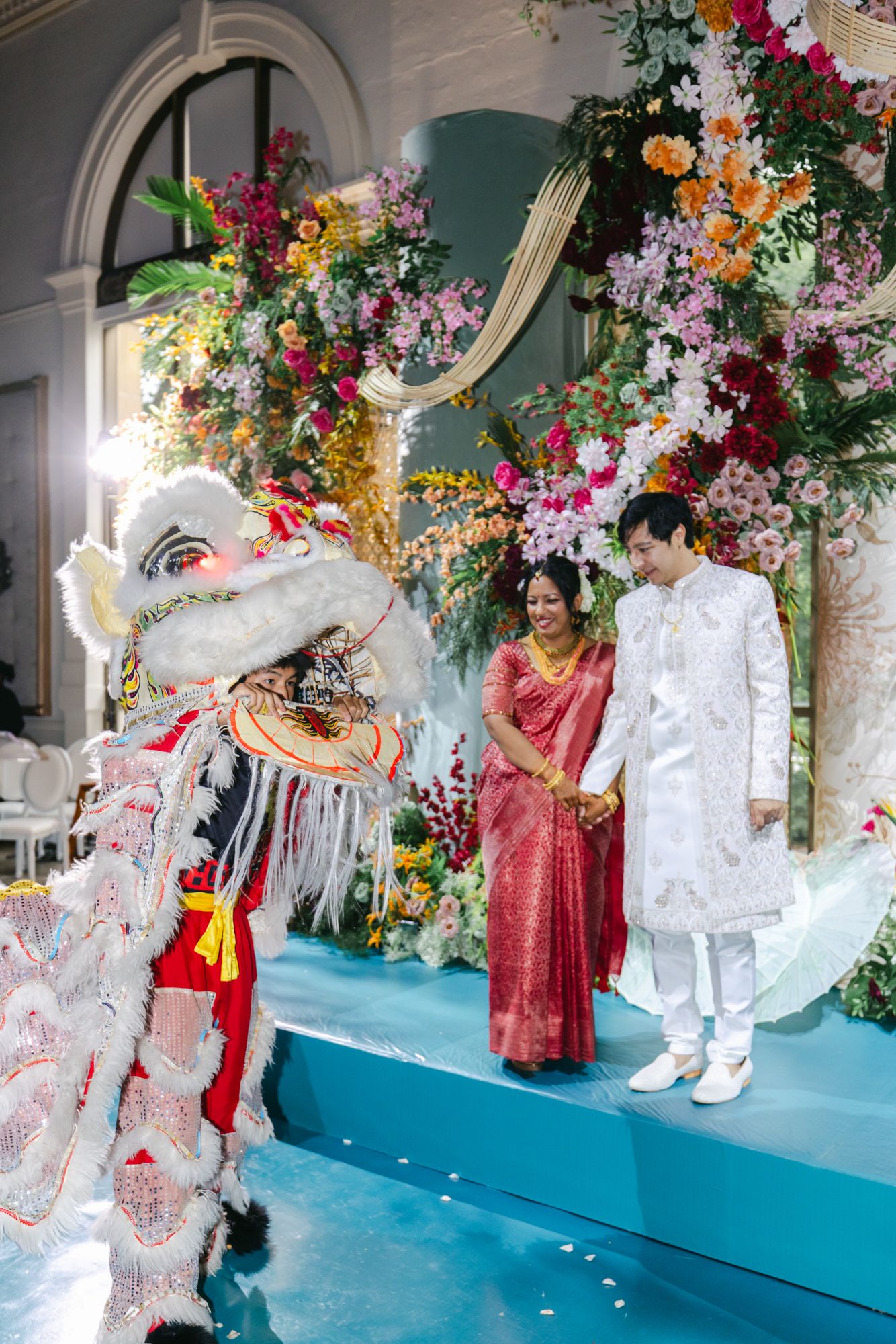 A bride in a red sari and a groom in a white sherwani smile beside a traditional lion dance performer at their wedding.