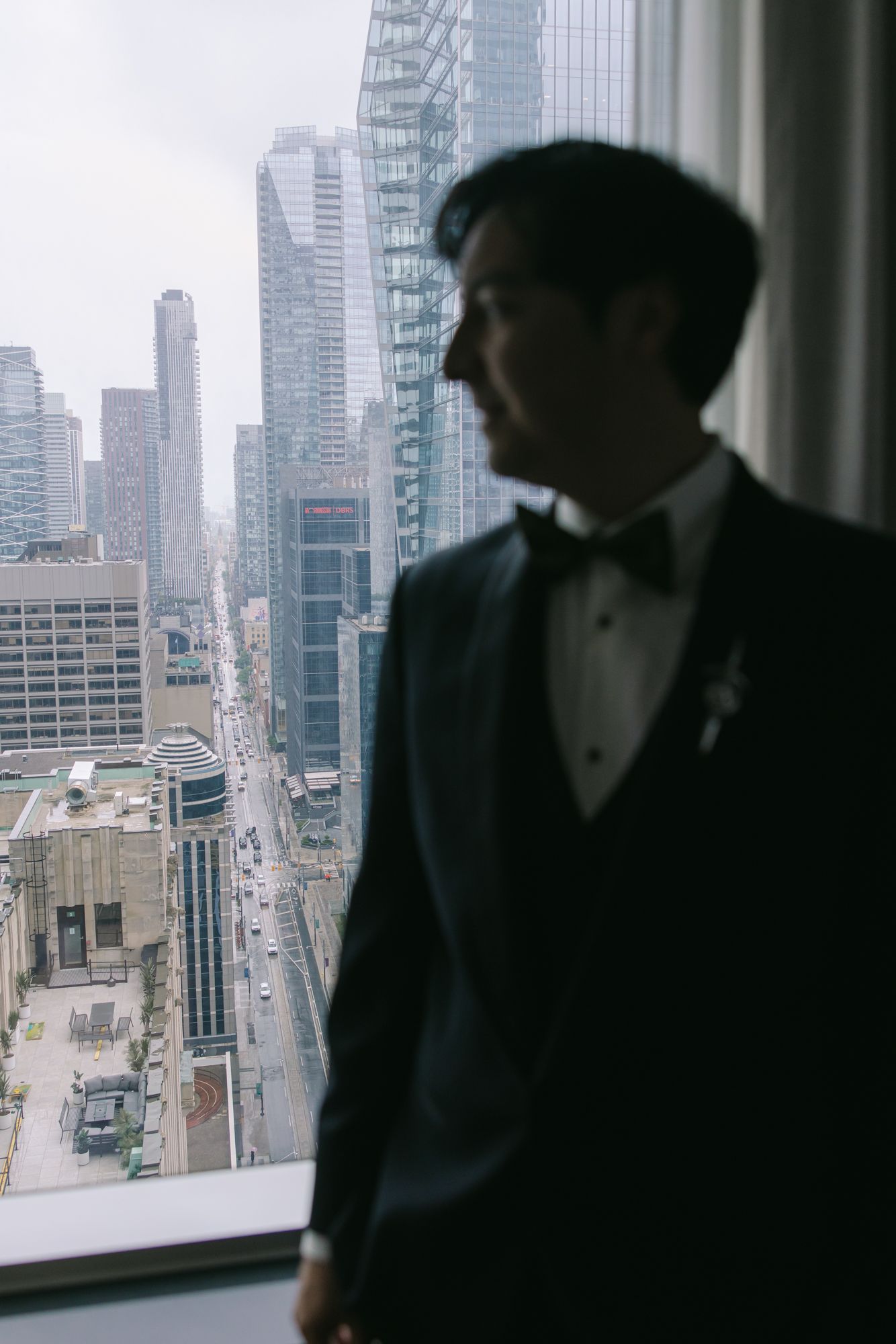 Silhouette of a groom in a dark suit and bow tie gazing out a window at a hazy city skyline with tall skyscrapers.