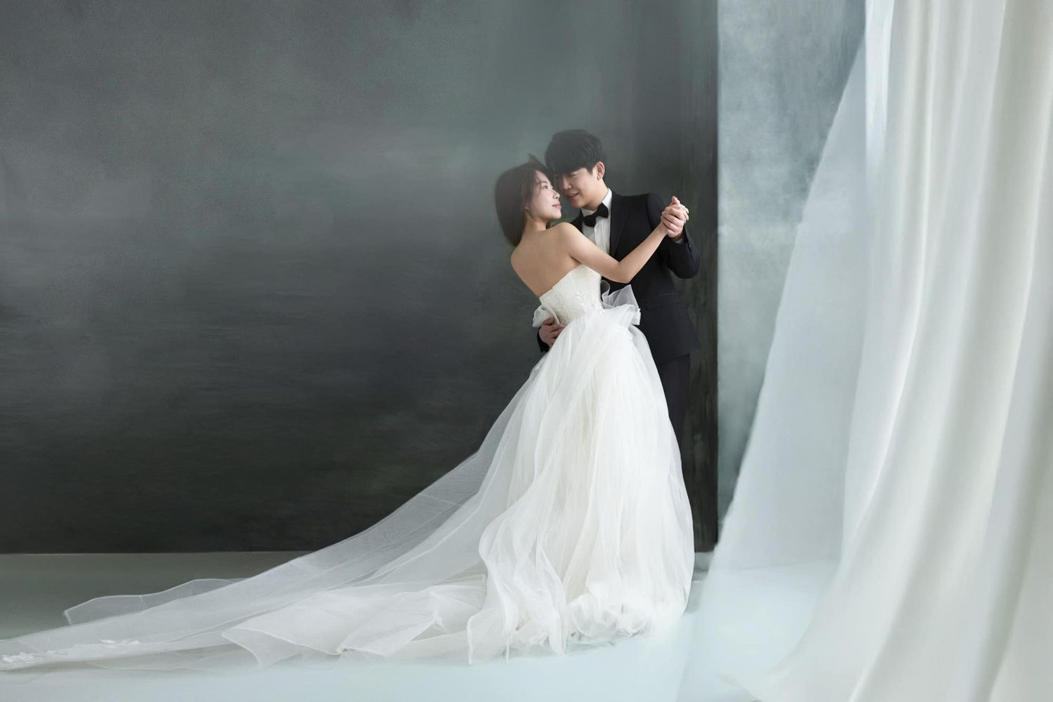 Couple sharing romantic dance in flowing white wedding gown against dramatic dark studio backdrop.