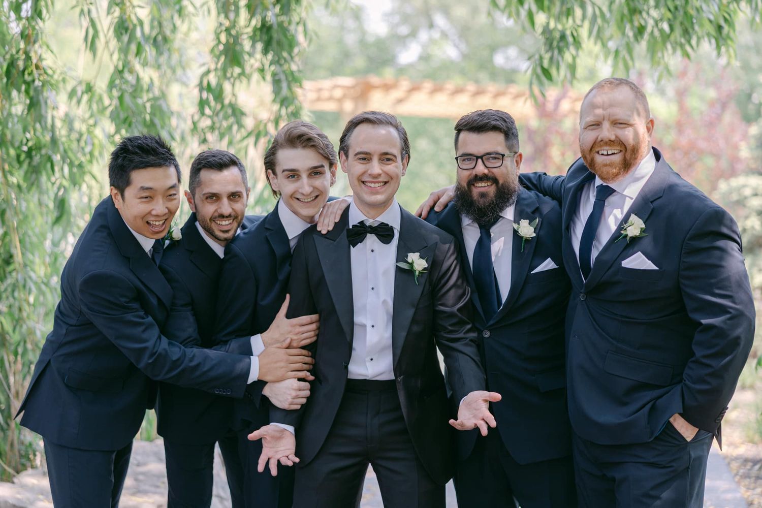 Groom celebrating with groomsmen in navy tuxedos under green trees at outdoor wedding.