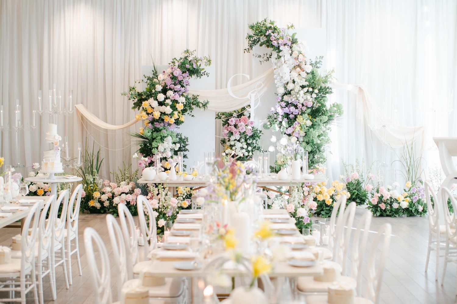 Stunning wedding reception hall with floral arch, pastel blooms, white ghost chairs, and draped ceiling decor.