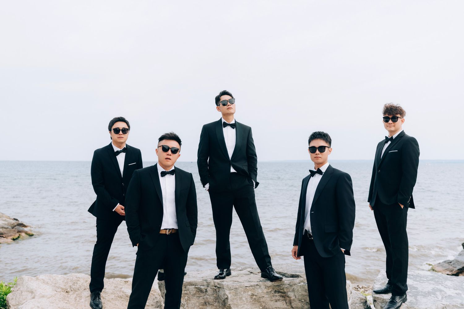 Five groomsmen in black tuxedos and sunglasses pose by the ocean.