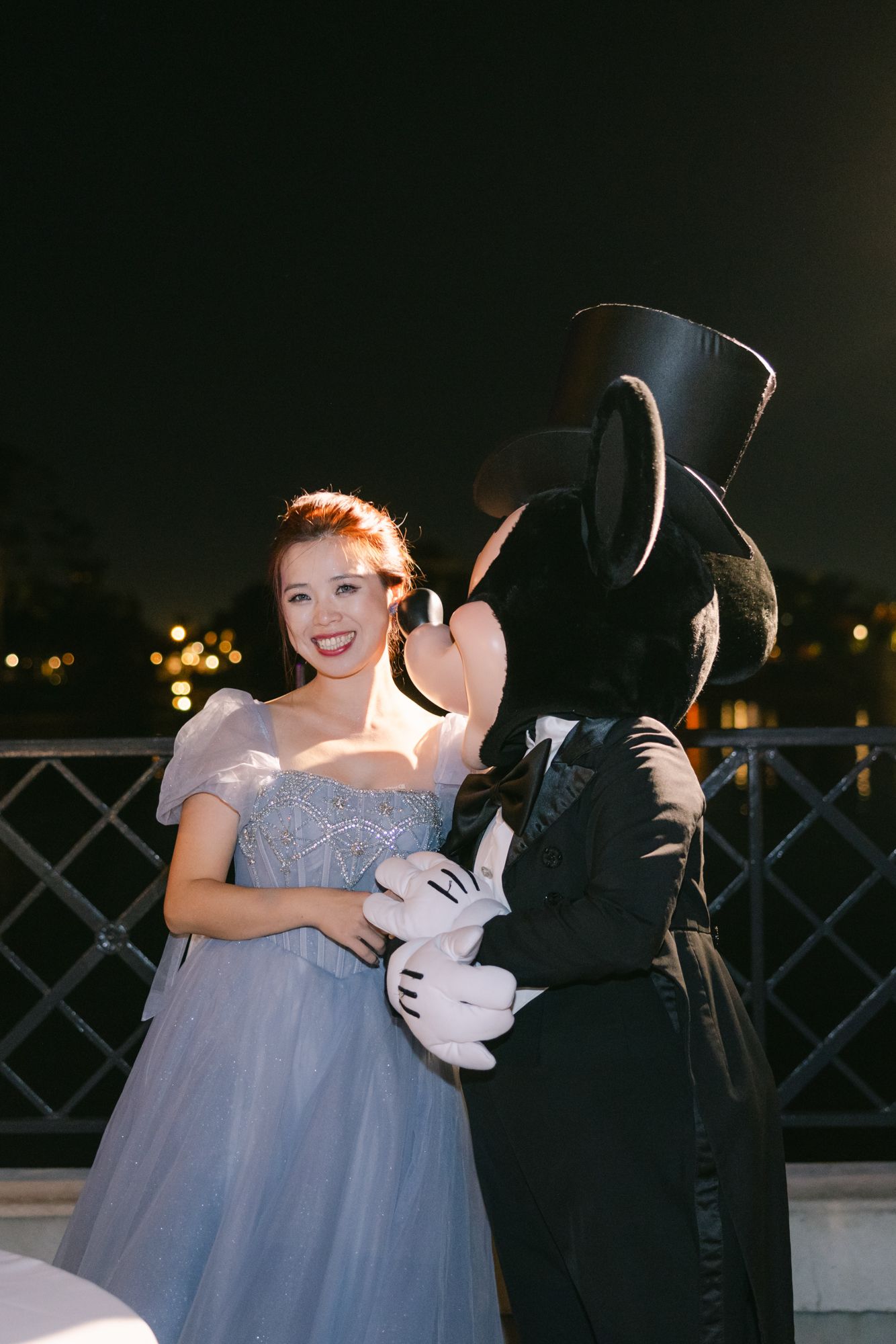 Smiling bride in blue gown poses with Mickey Mouse in tuxedo at Disney World at night.