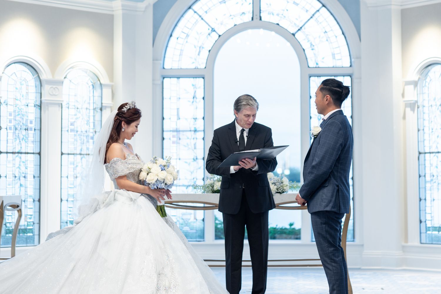 Bride and groom exchange vows before an officiant inside a bright, elegant chapel with arched windows.