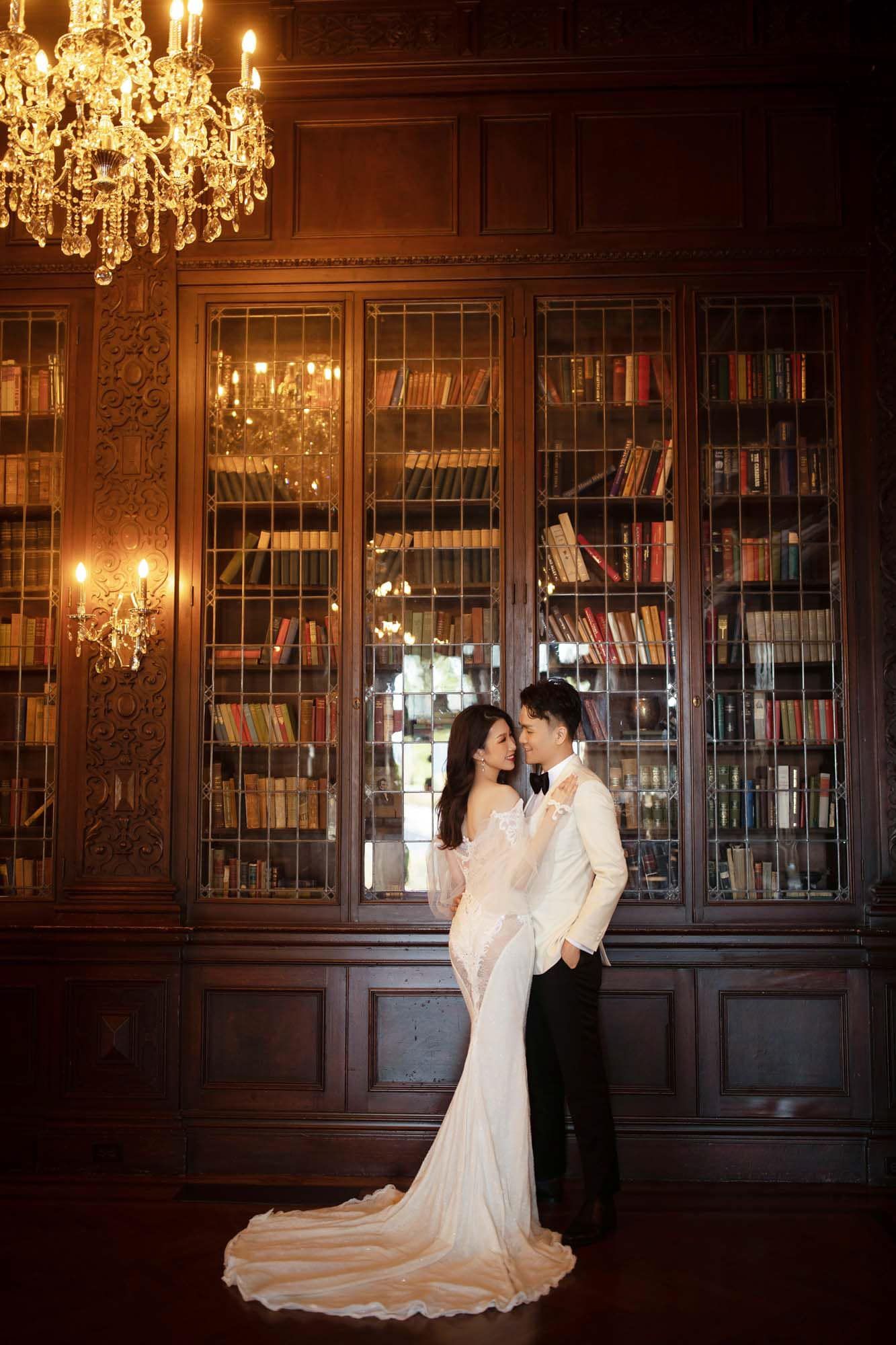 Elegant wedding couple embracing in a grand library with ornate bookshelves and a glowing crystal chandelier.