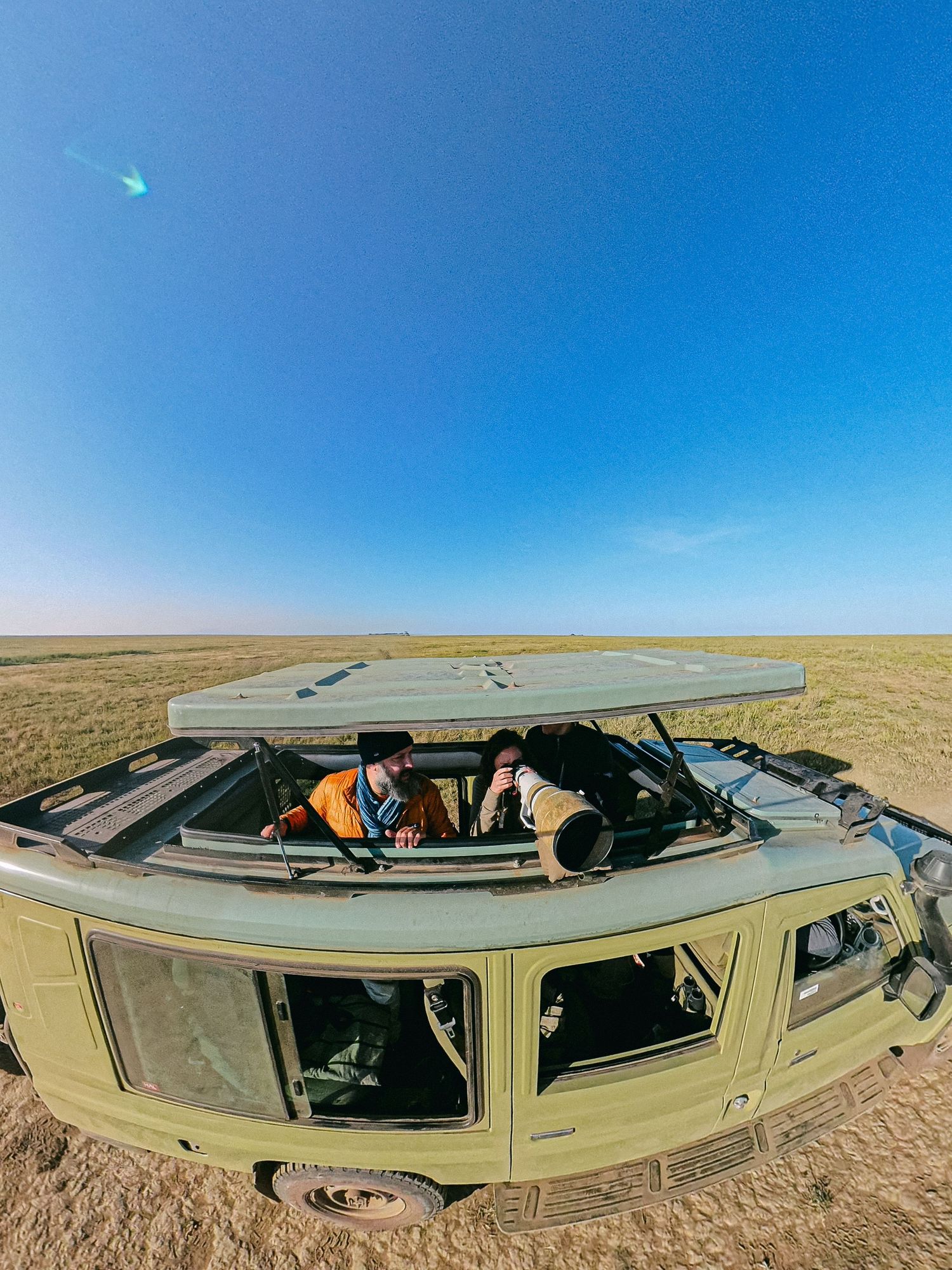 Two photographers shooting from the open roof hatch of a vintage green van on vast open grasslands under a clear blue sky.