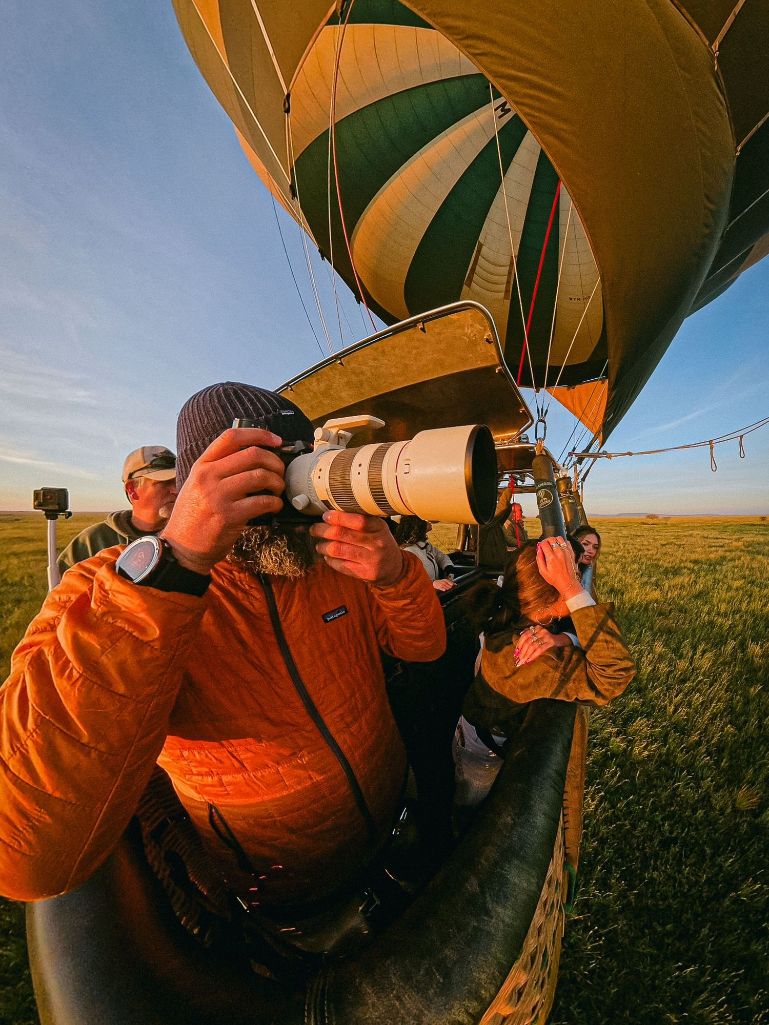 Photographer in orange jacket uses telephoto lens near a green and gold hot air balloon at sunrise in a field.