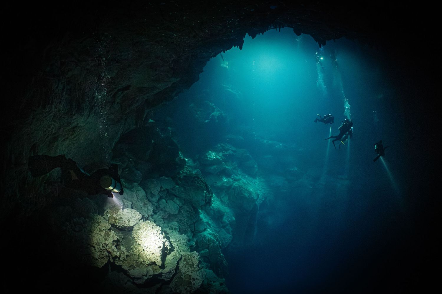 Underwater cave divers explore dark caverns illuminated by blue light beams penetrating through crystal clear water.