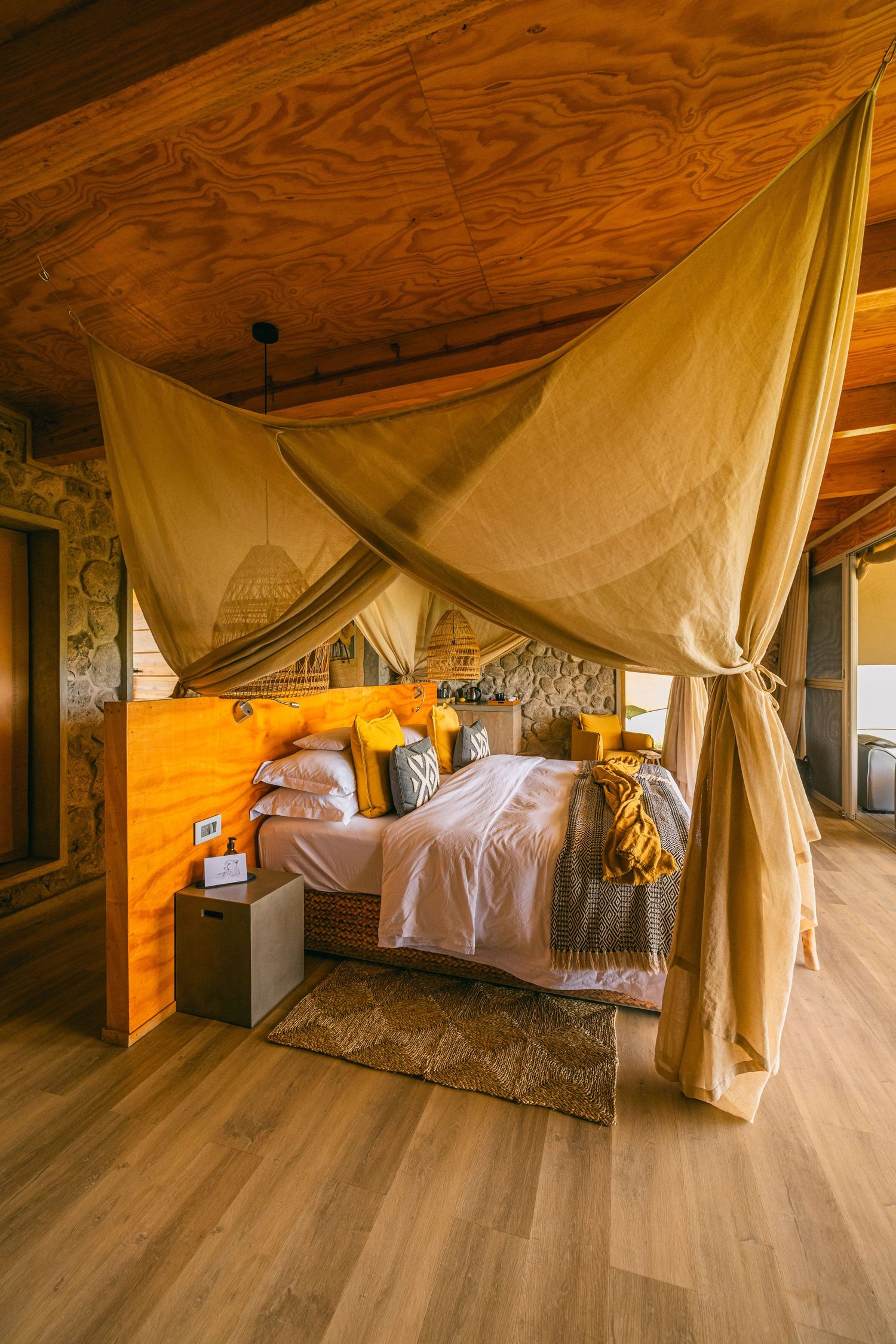 Rustic bedroom with canopy bed, wood ceiling, stone walls, yellow accents, and warm lighting in a cozy lodge setting.