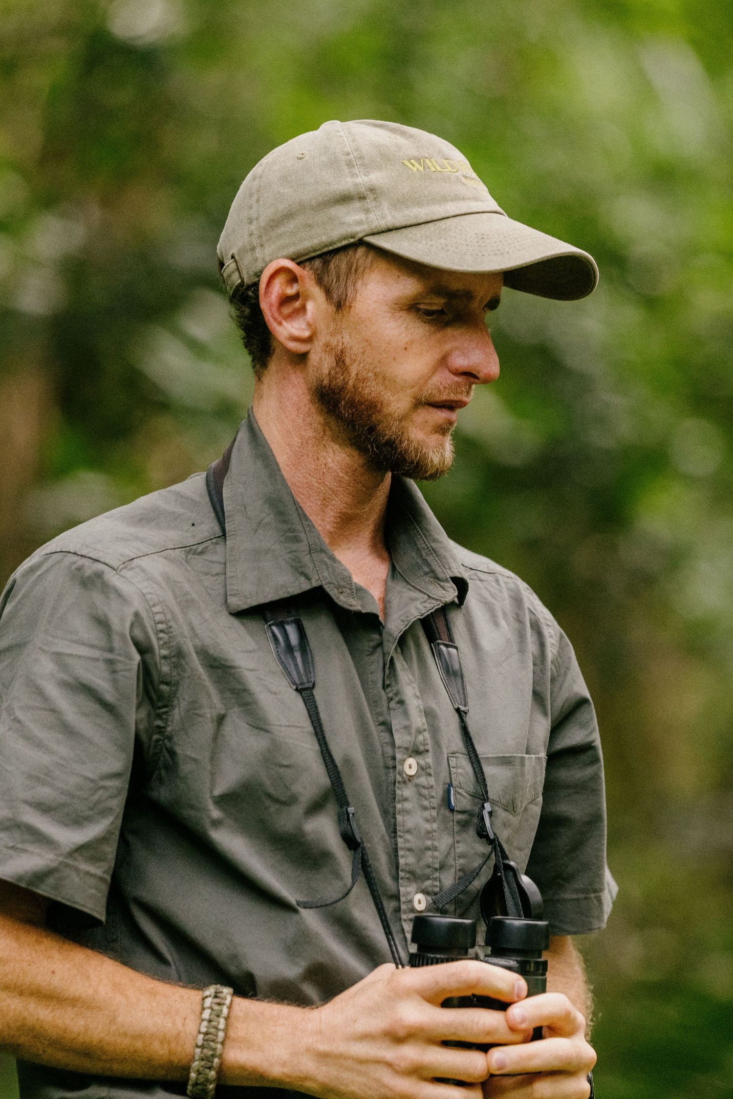 Person wearing a gray shirt and baseball cap examines a handheld device outdoors against a blurred green background.