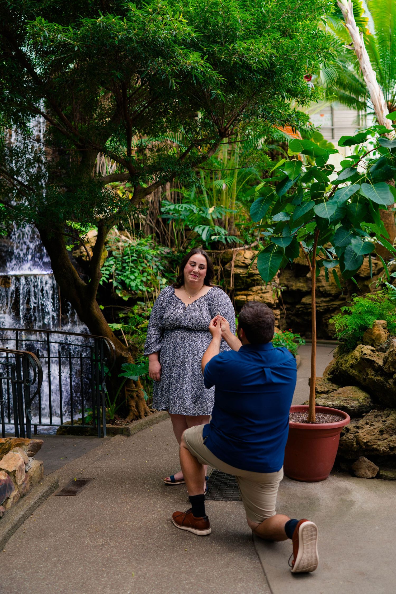 Man on one knee proposing to woman near a waterfall in a tropical garden.