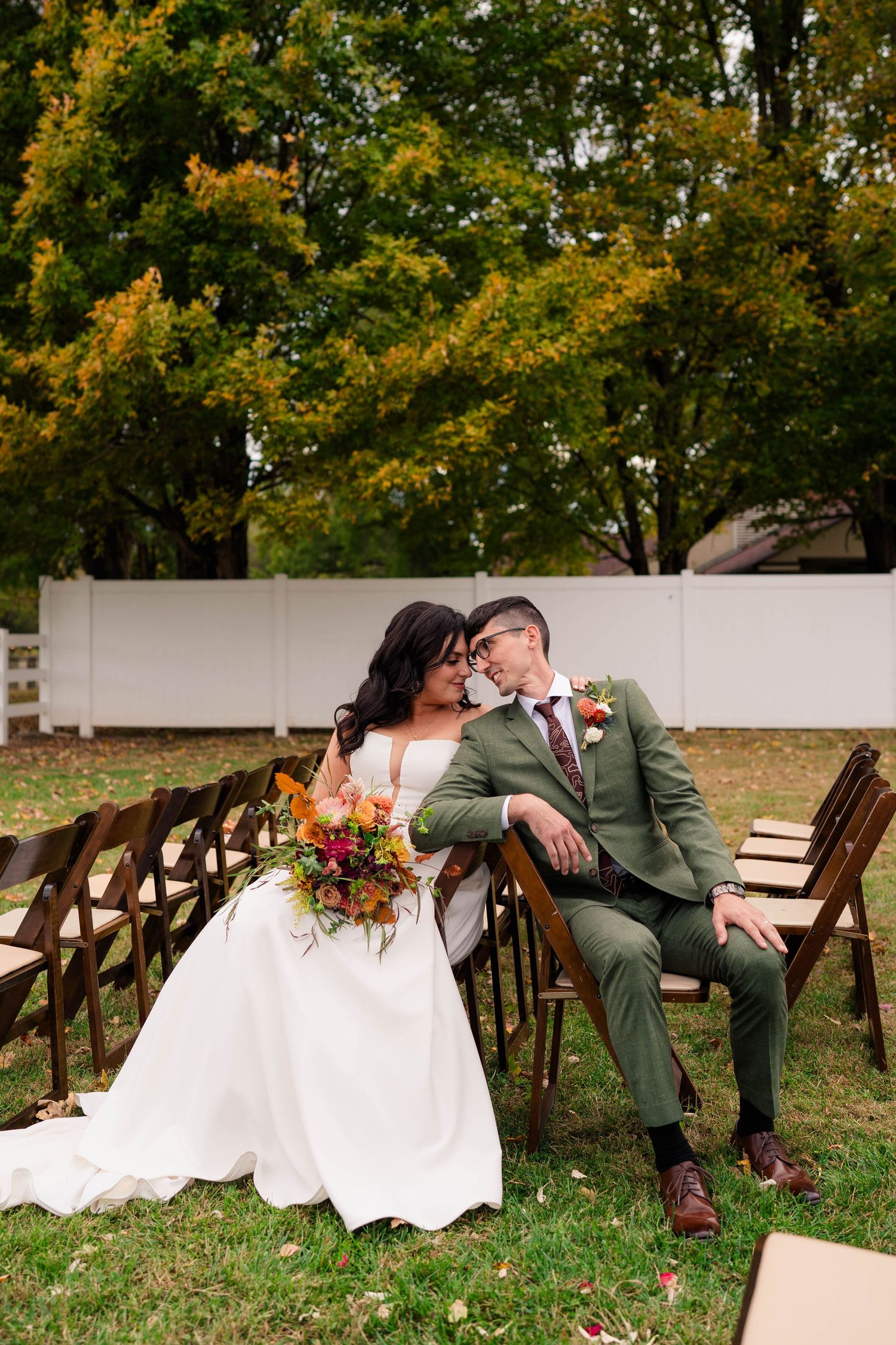 Bride and groom sit closely together in wooden chairs at outdoor fall wedding ceremony venue.