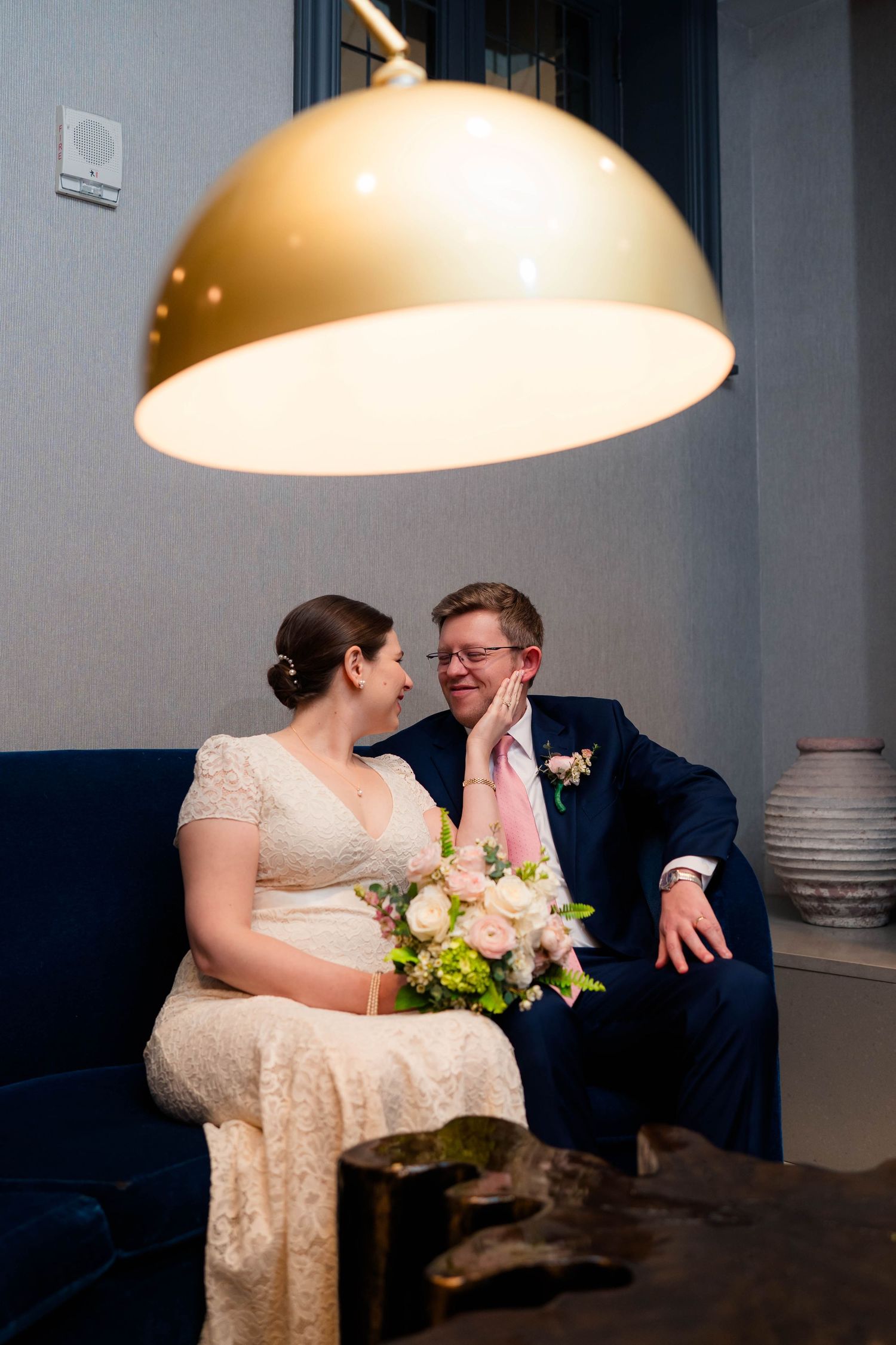 Bride and groom sharing a tender moment on a blue velvet sofa under a warm gold pendant light.