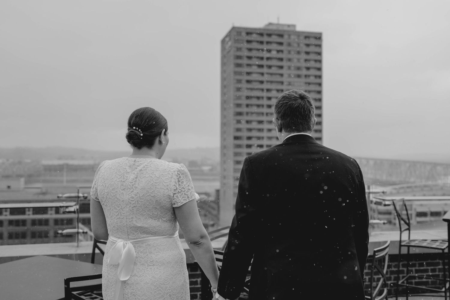 Black and white photo of bride and groom holding hands while gazing at a rainy city skyline together.