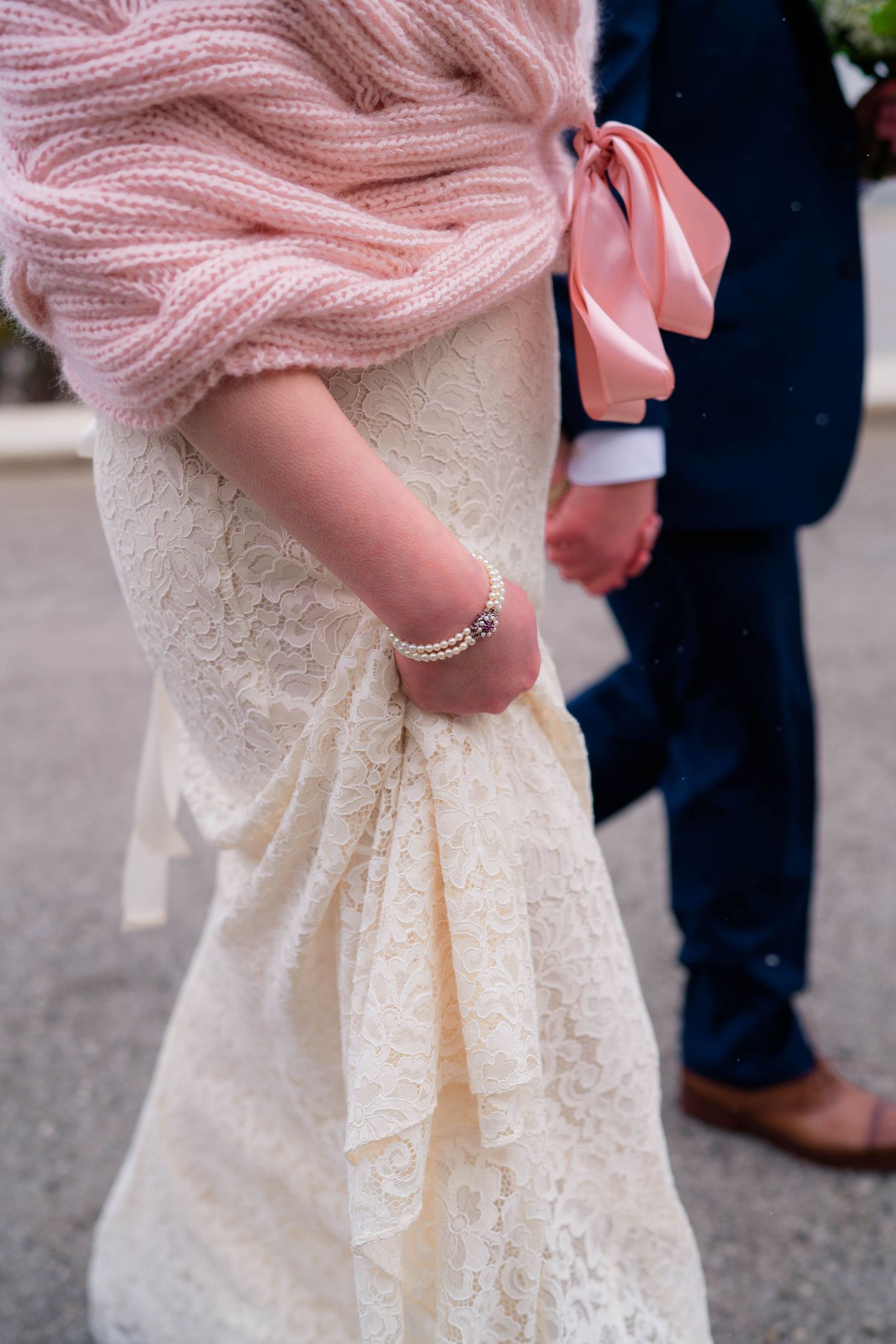 Close-up of bride in lace wedding gown and pink knit shawl holding grooms hand with pearl bracelet visible.