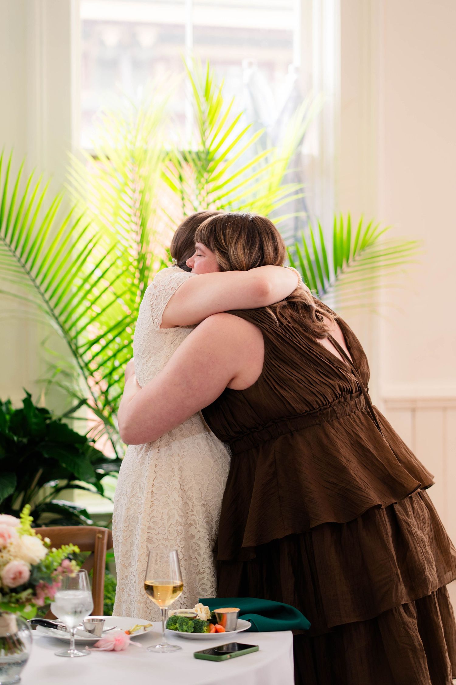 Bride in lace dress and woman in brown ruffled dress sharing a warm emotional hug at the reception.