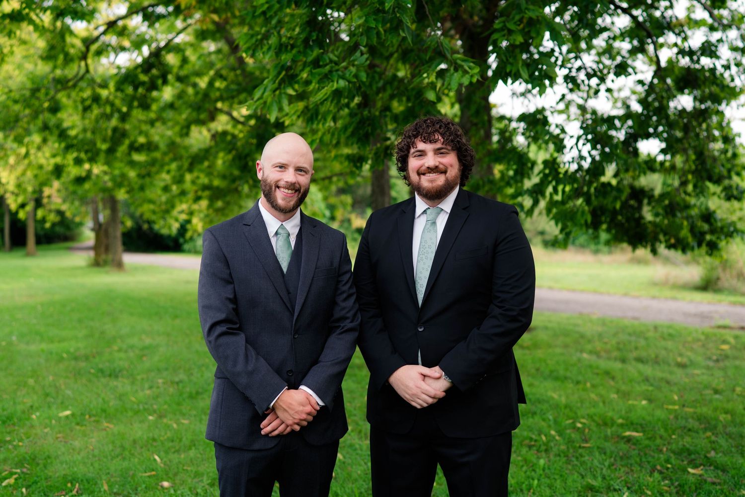 Two groomsmen in black suits smiling together outdoors under a large tree.