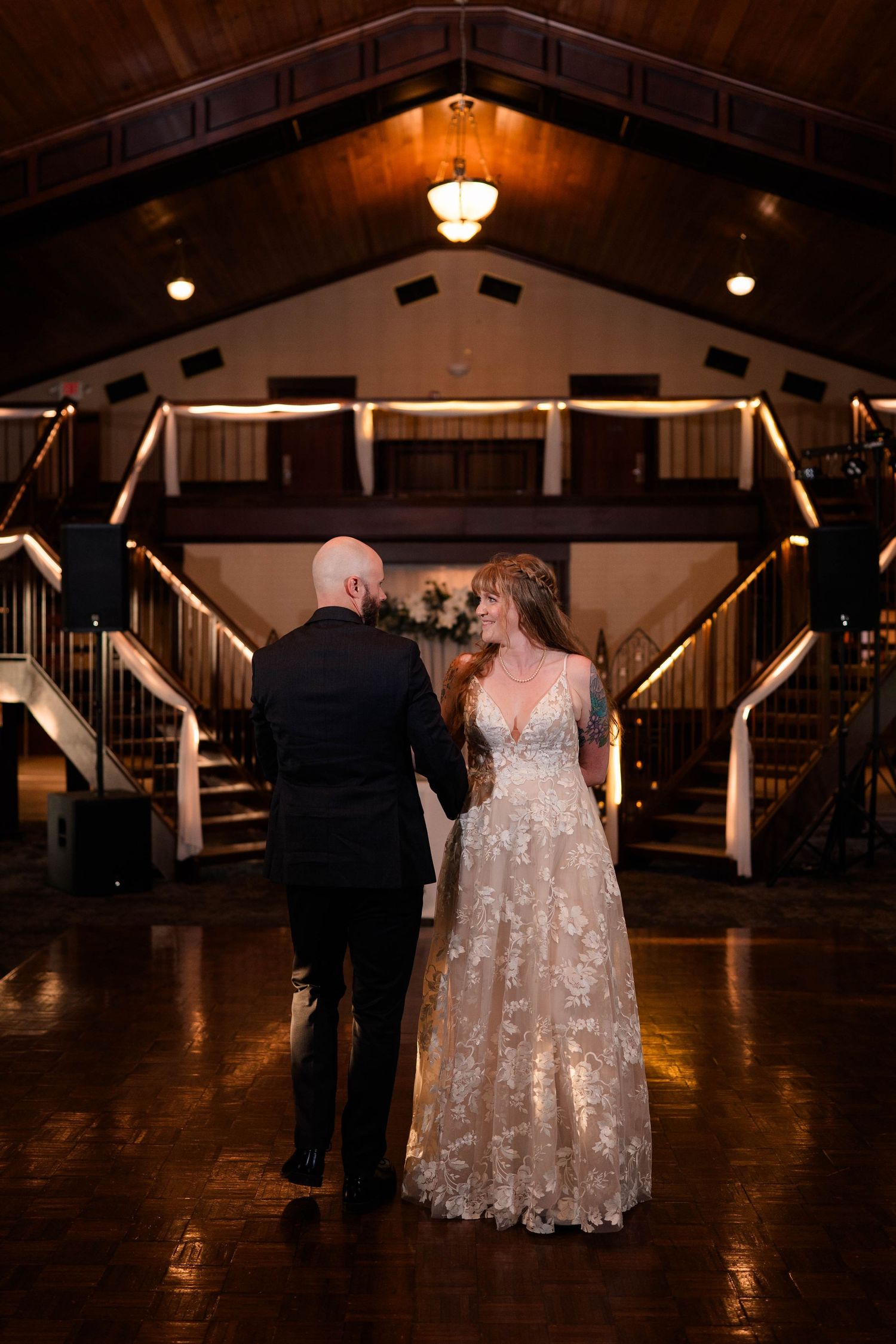 Bride and groom share first dance on wooden dance floor beneath warm cathedral ceiling lights at wedding reception.