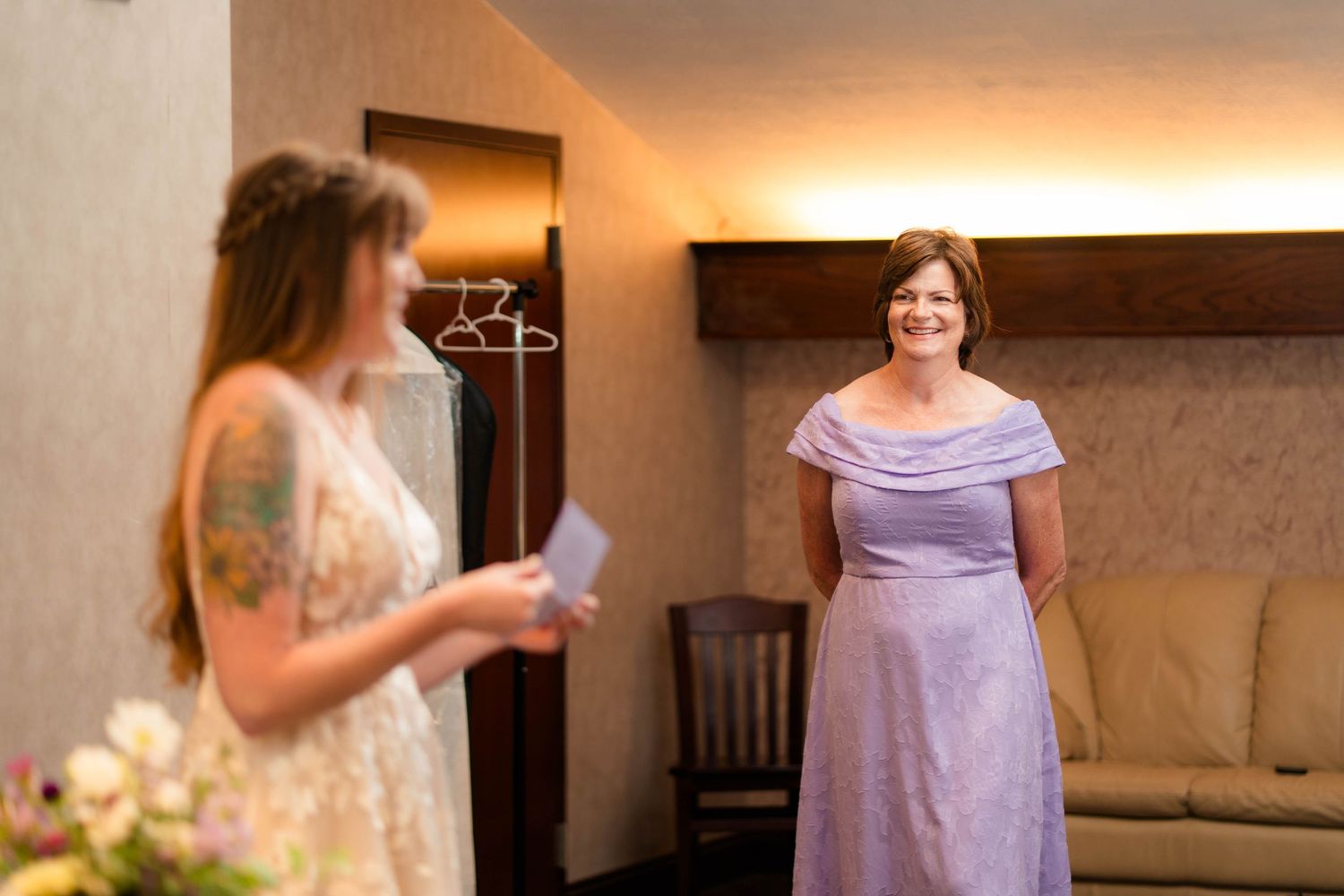 A bride in a white lace gown shares a joyful moment with a woman in a lavender dress inside a hotel room.