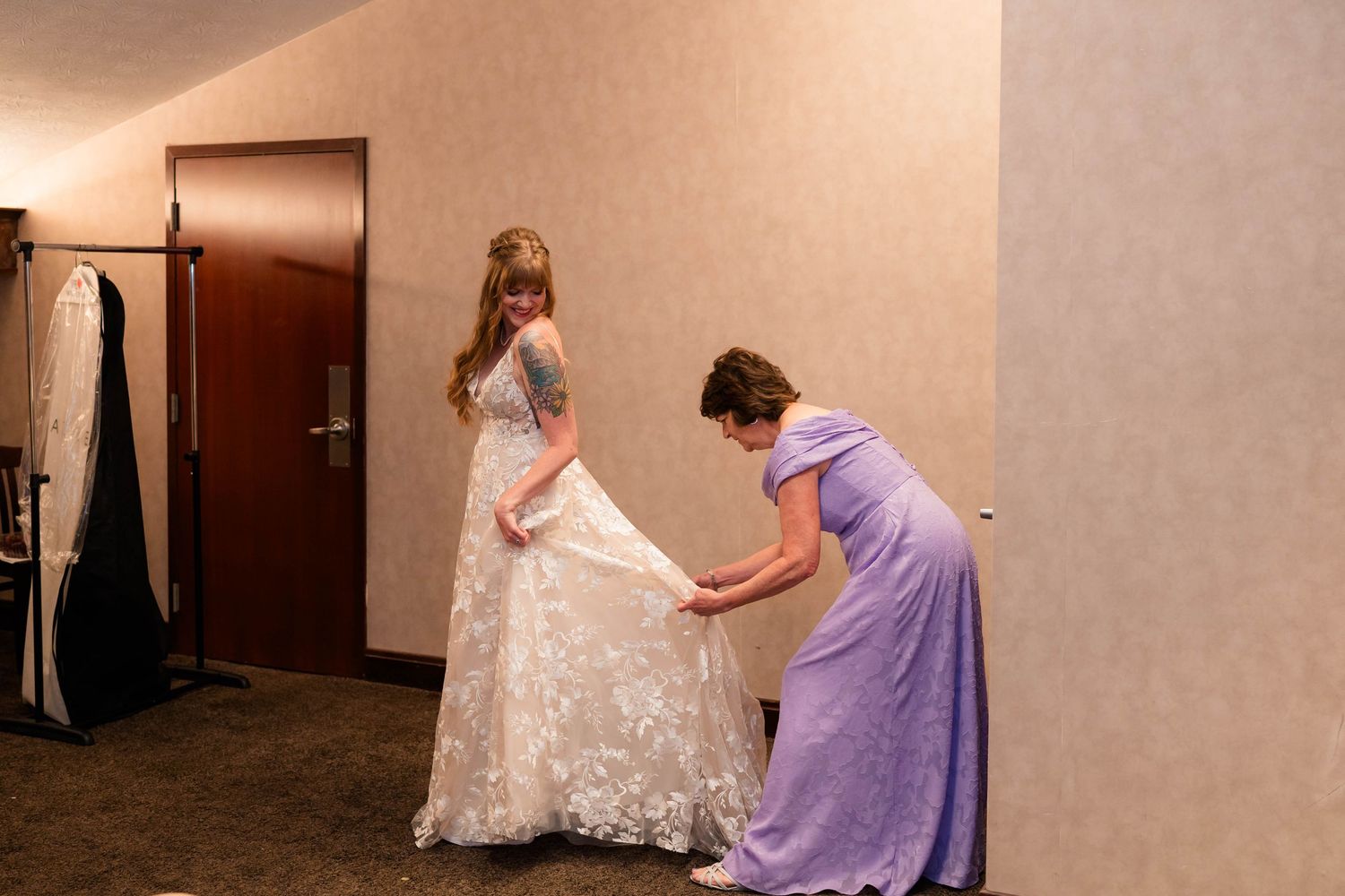 Woman in white lace wedding gown being helped by bridesmaid in purple dress in hotel corridor.