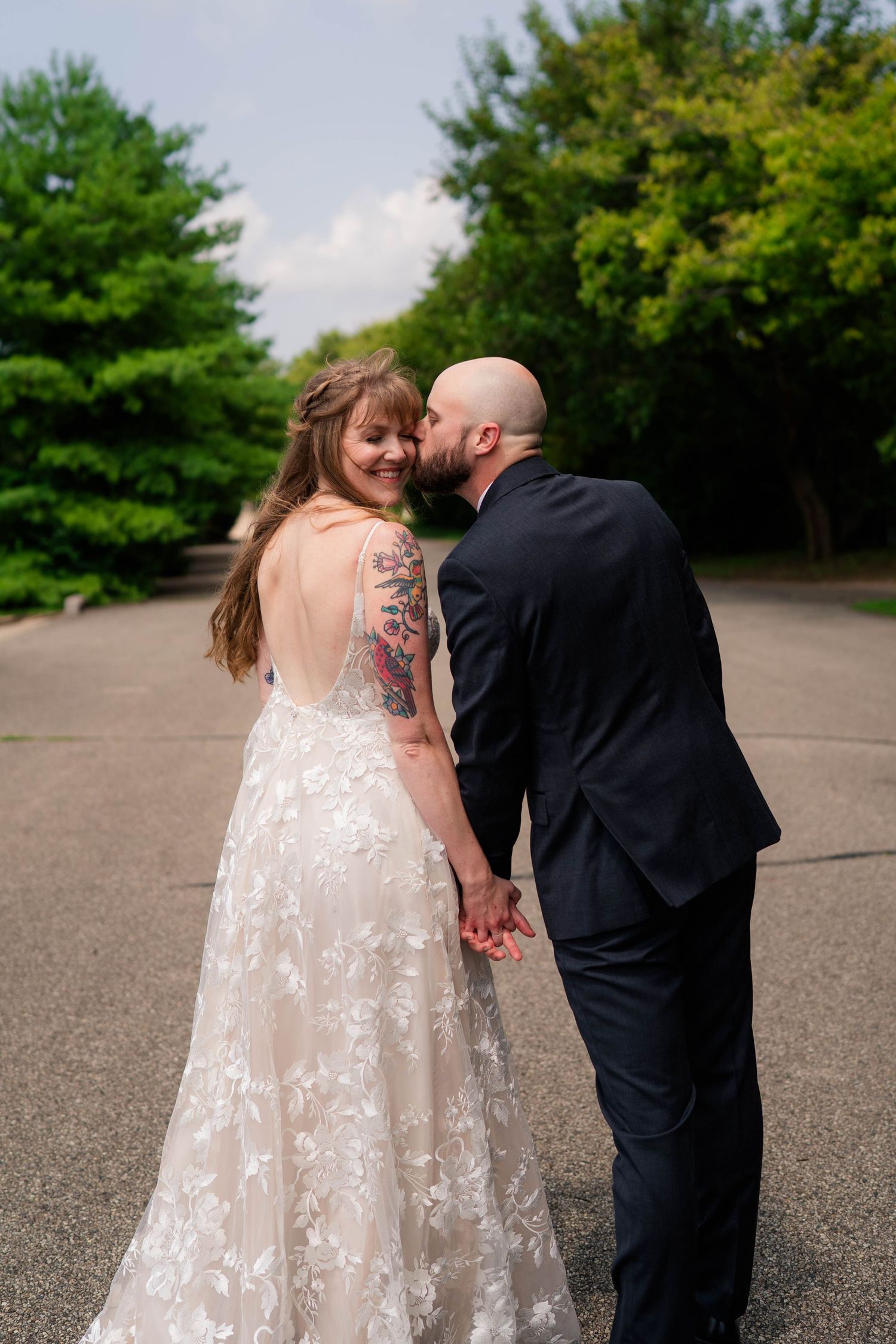Bald groom kissing tattooed bride on cheek as she smiles over shoulder in backless lace wedding gown outdoors.