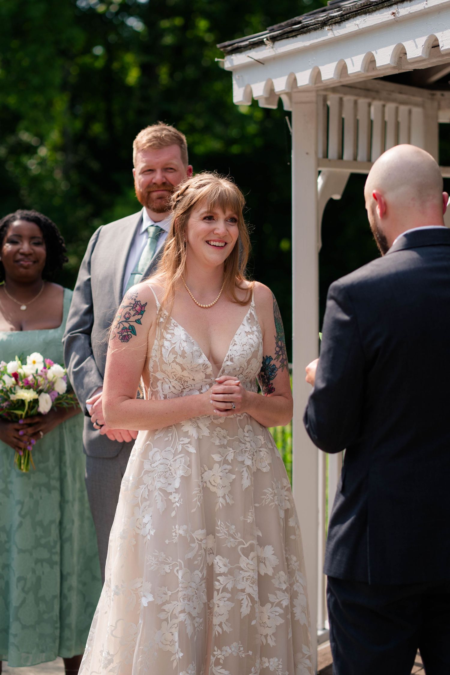 Smiling bride in lace gown listens to groom read vows at an outdoor wedding ceremony.