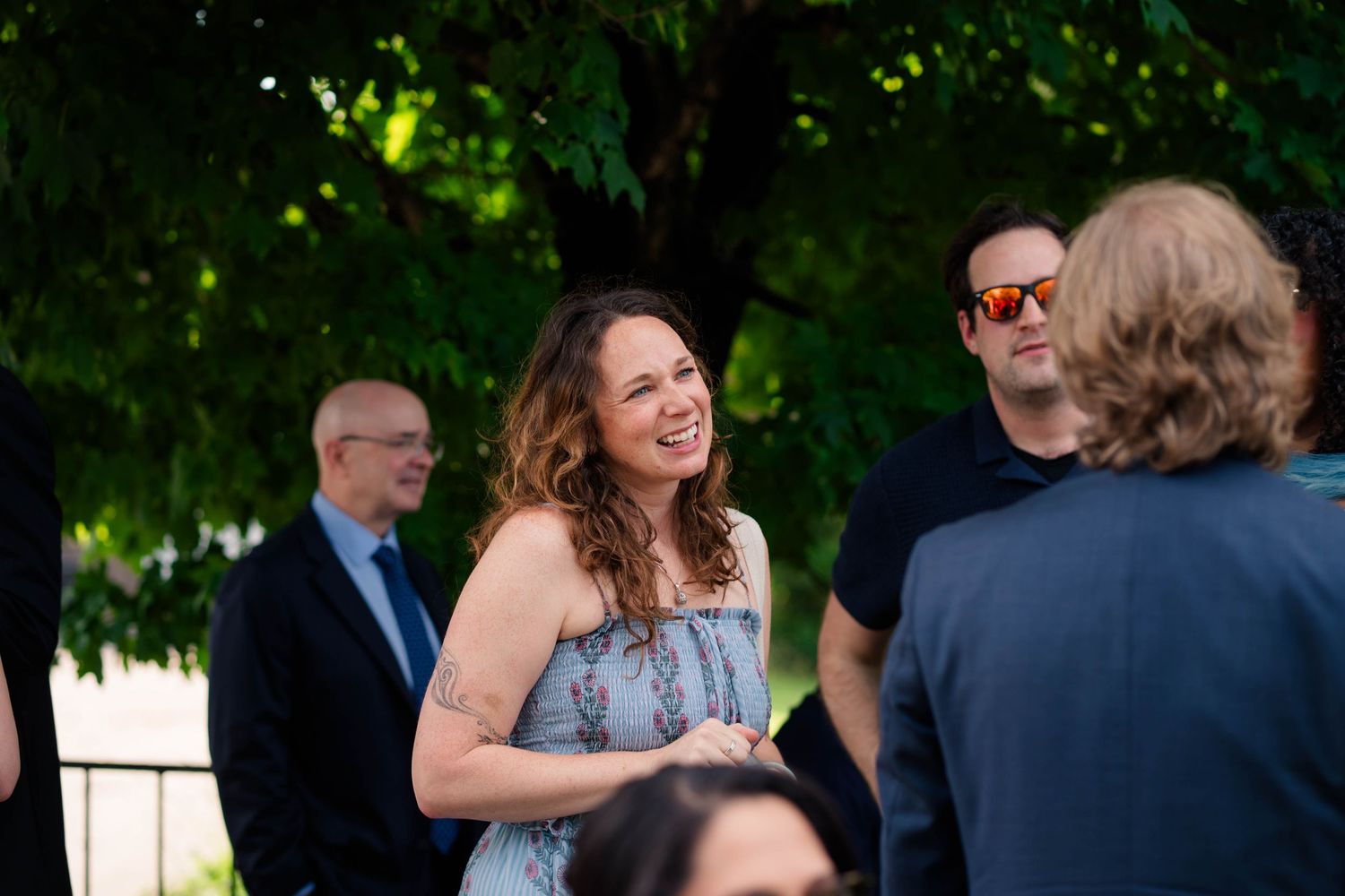 A woman laughs joyfully at an outdoor wedding reception surrounded by guests beneath green trees.