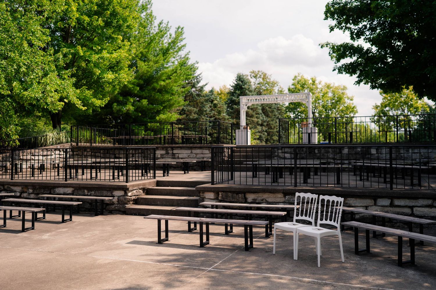 Outdoor wedding ceremony venue with wooden benches, white arbor, and lush green trees.