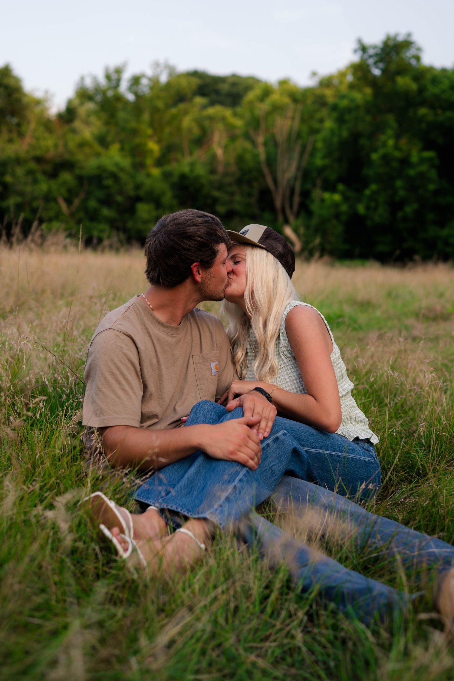 A couple shares a kiss while sitting in a green meadow, woman in a hat and casual jeans outfit.