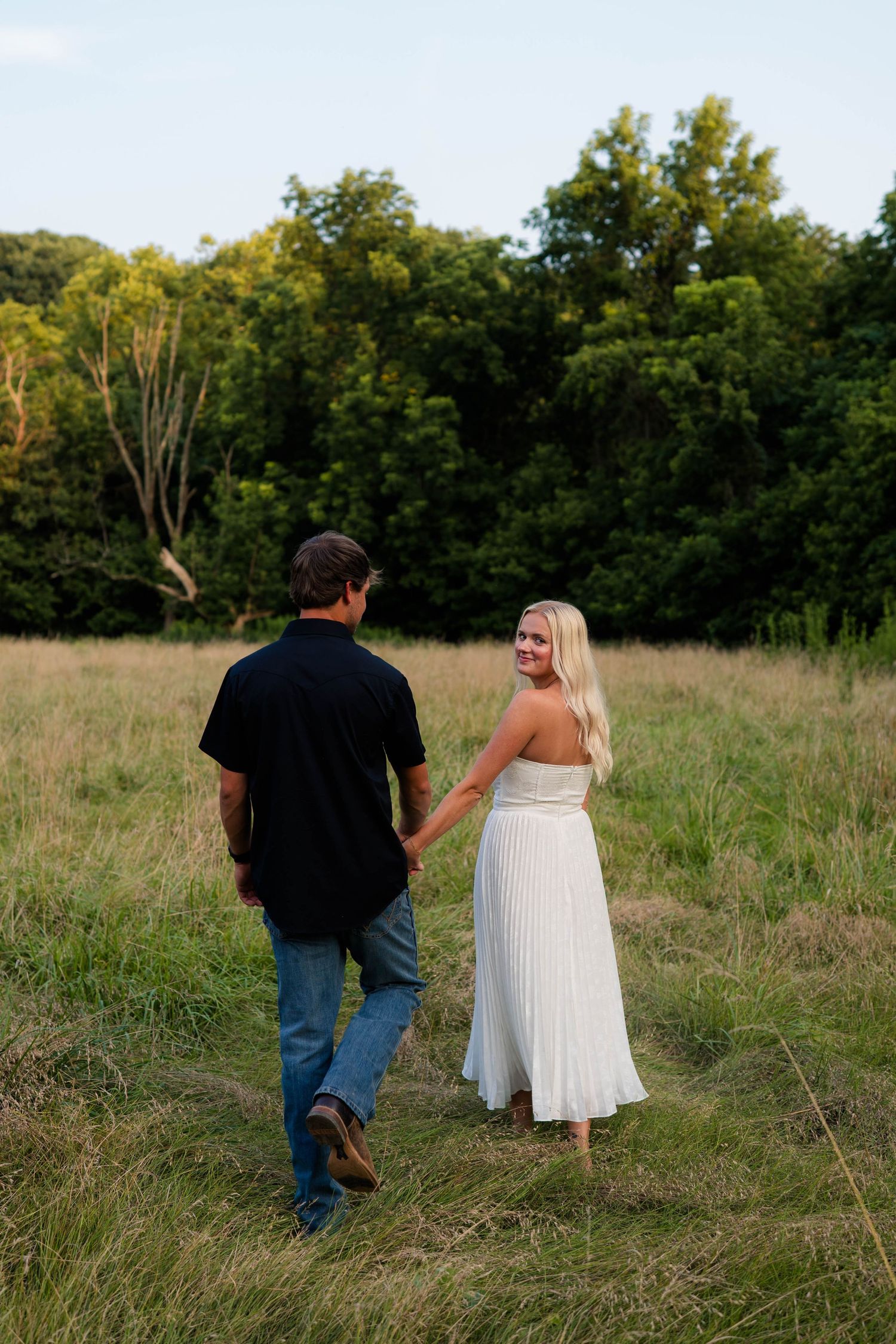 Couple holding hands walking through a lush green field at dusk, woman in white dress looking back.