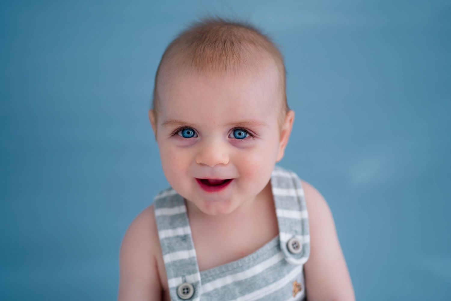 A cheerful baby with bright blue eyes smiles wearing a striped overalls romper against a blue backdrop.