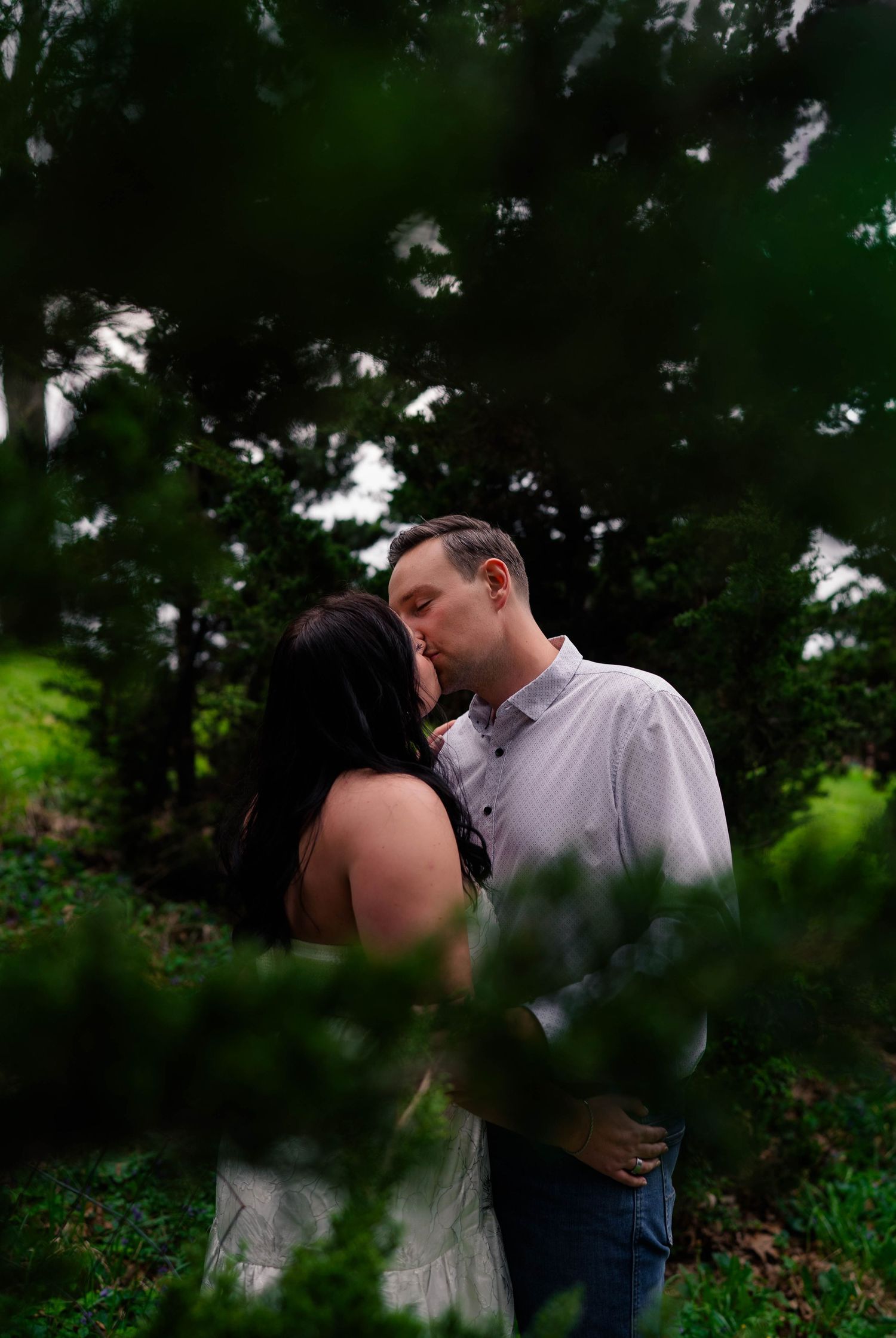 A couple shares a tender kiss framed by lush green foliage in a serene wooded setting.