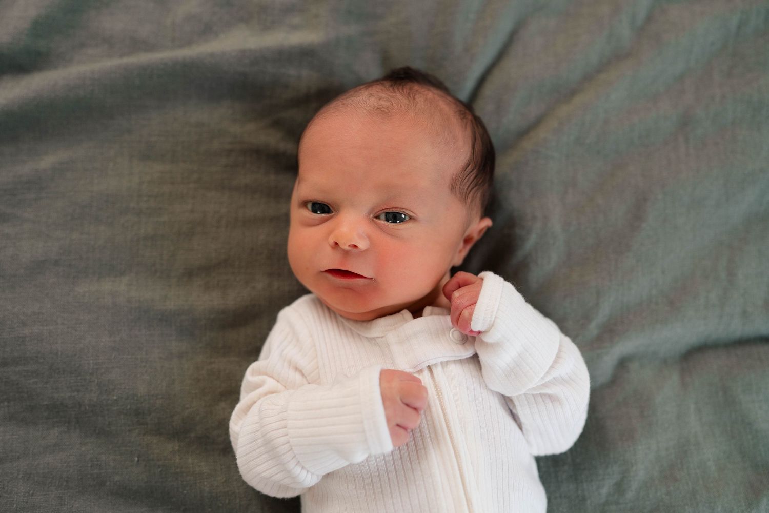 Alert newborn baby lying on a gray blanket wearing a white onesie, looking at the camera.