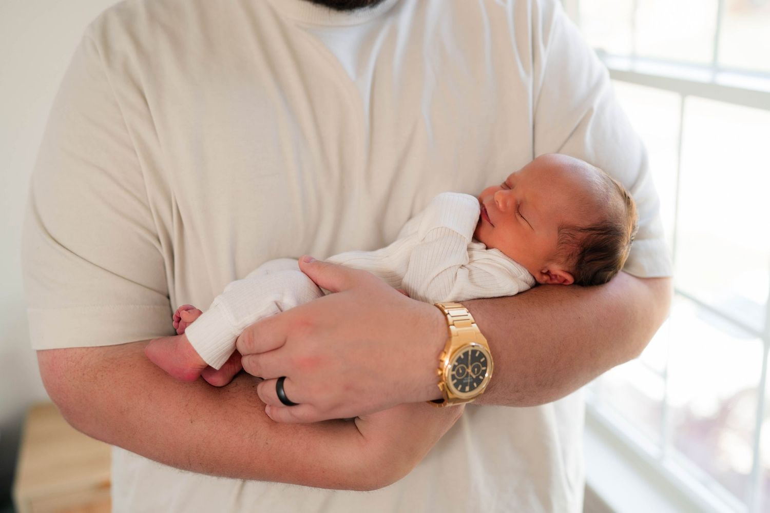 A father in a white shirt gently cradling a sleeping newborn baby in his arms, wearing a gold watch.