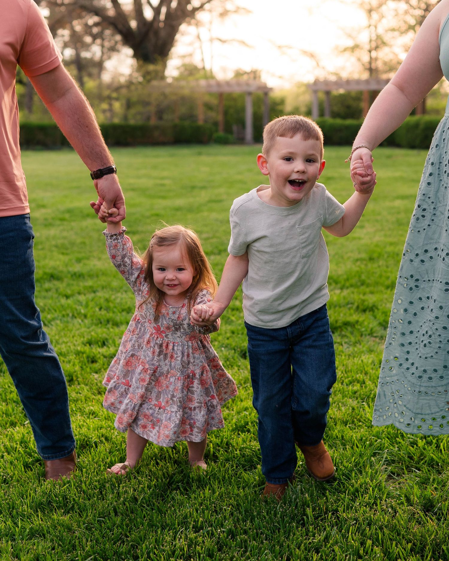 Two young children holding hands with parents on green grass at sunset in a joyful outdoor family photo.