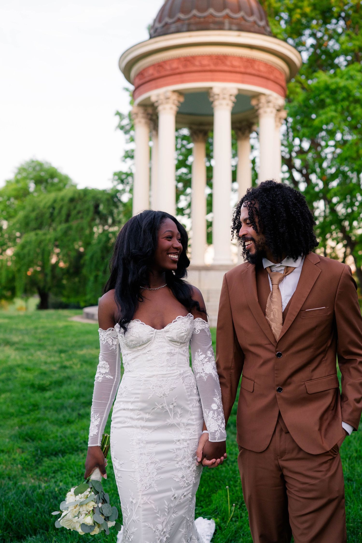 Joyful Black bride in lace gown and groom in brown suit hold hands near elegant rotunda.