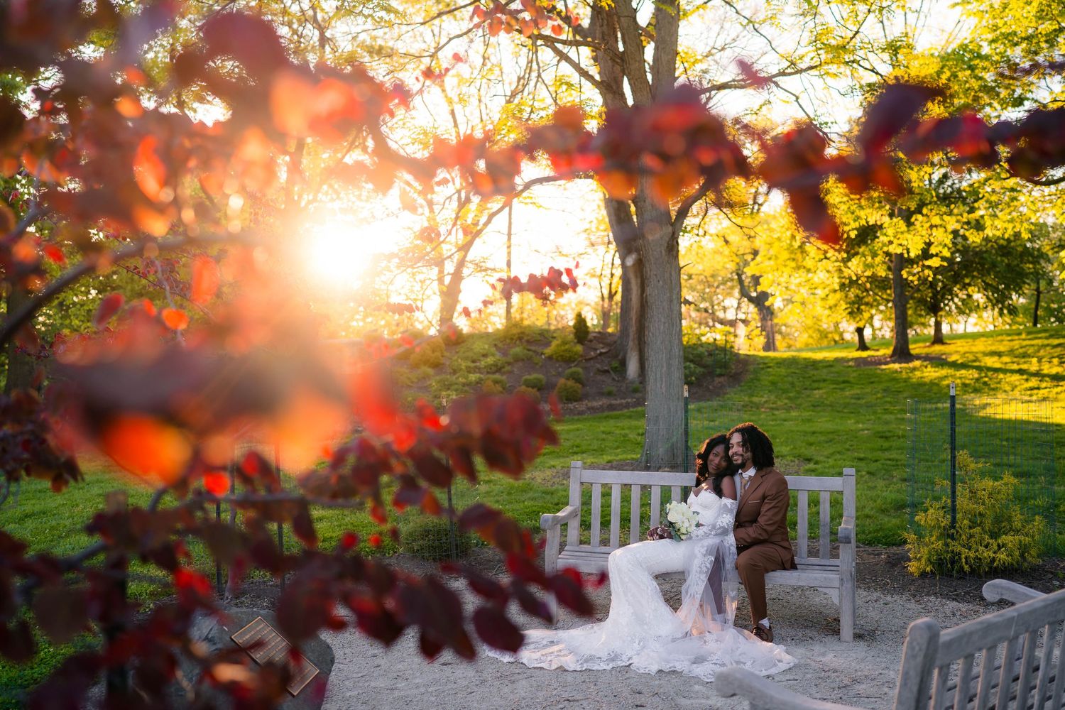 Bride and groom sitting on a white bench surrounded by colorful autumn foliage at golden hour.