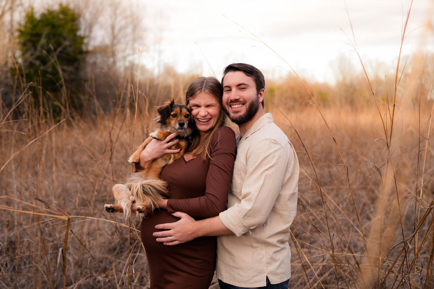 Happy couple embracing with their dog in a dry autumn field during a maternity photo shoot.