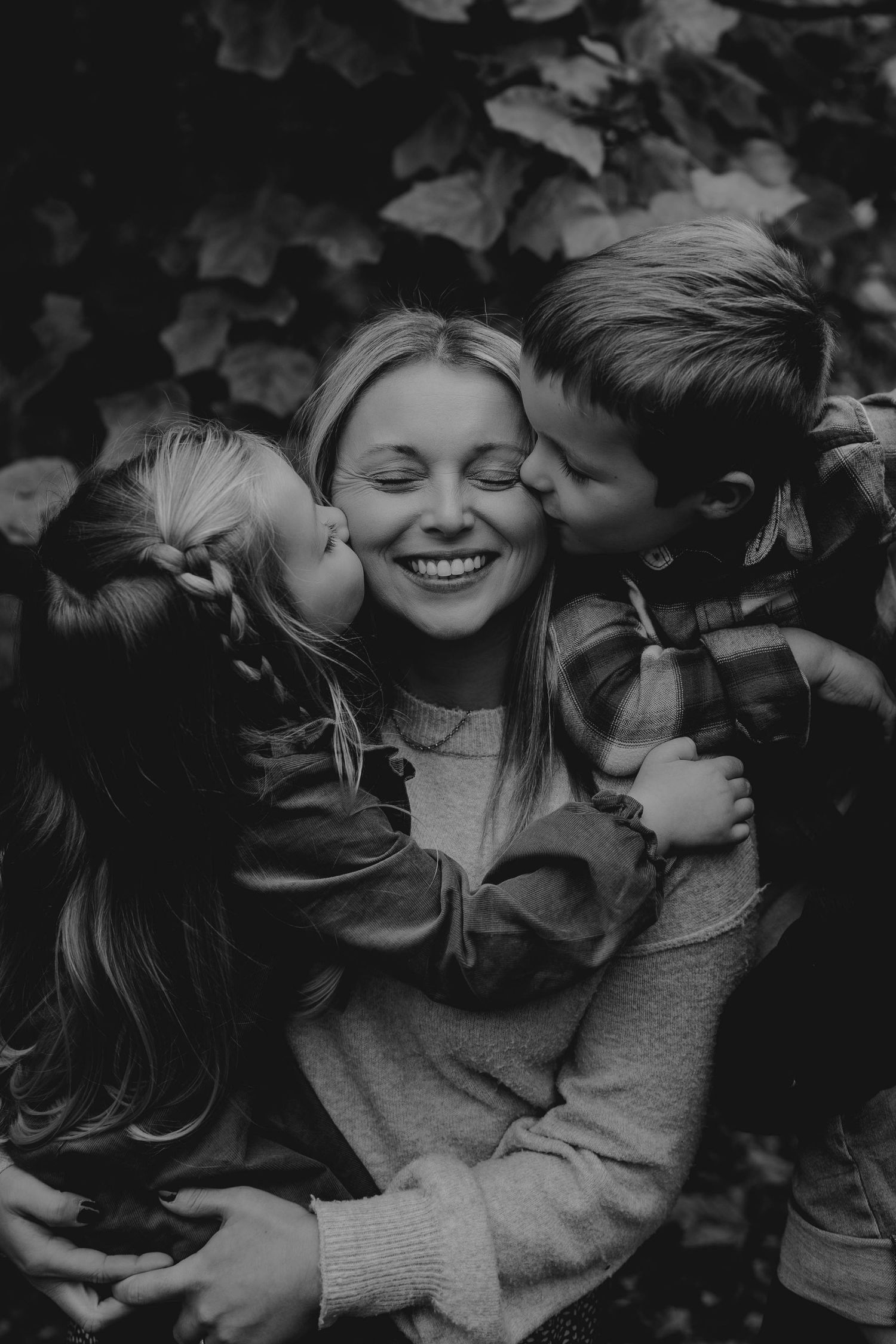 Black and white photo of a smiling mother being kissed on both cheeks by her two young children outdoors.