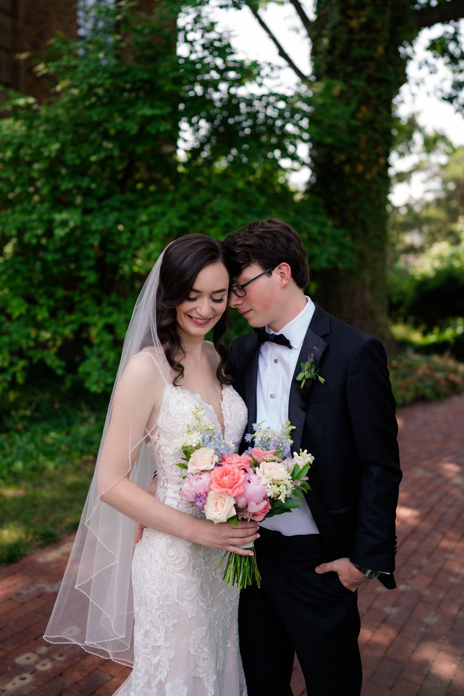Bride and groom share a tender moment outdoors, she holds a pink and blue bouquet, wearing lace gown and veil.