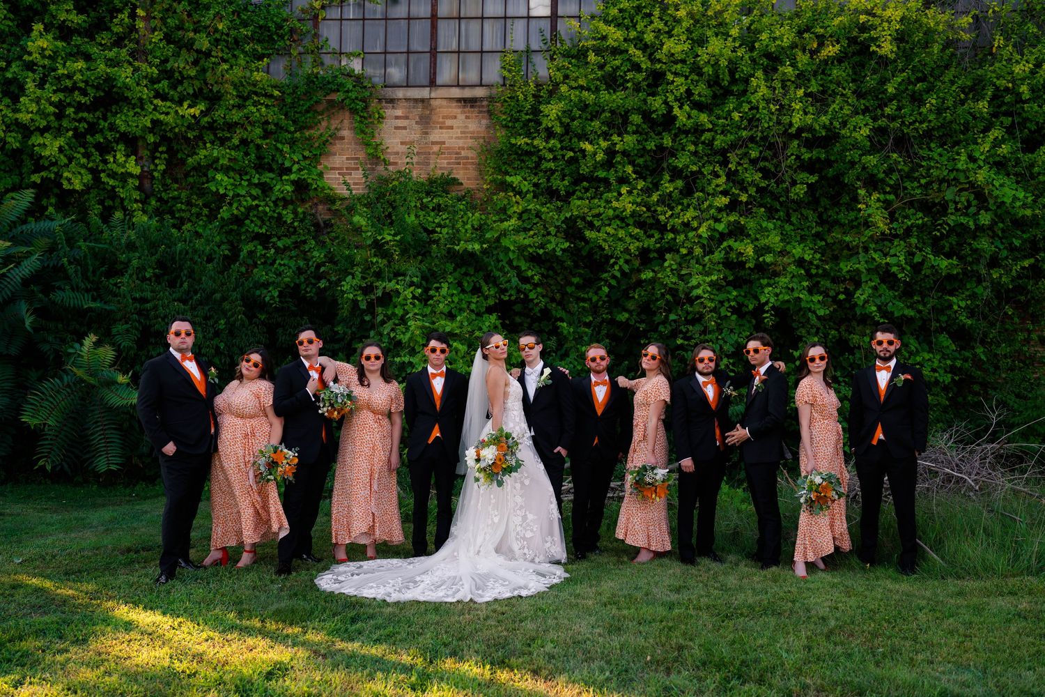 Wedding party poses outdoors in front of lush greenery, bridesmaids in blush gowns and groomsmen in black.