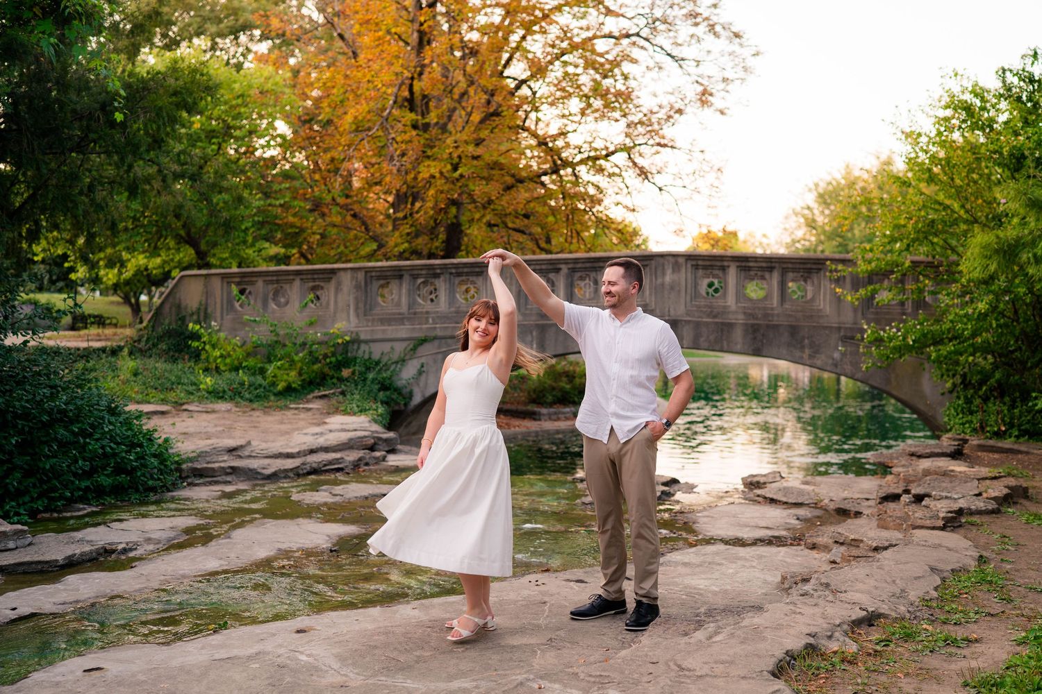 Couple dancing and twirling near a stone bridge by a pond in a scenic park at sunset.