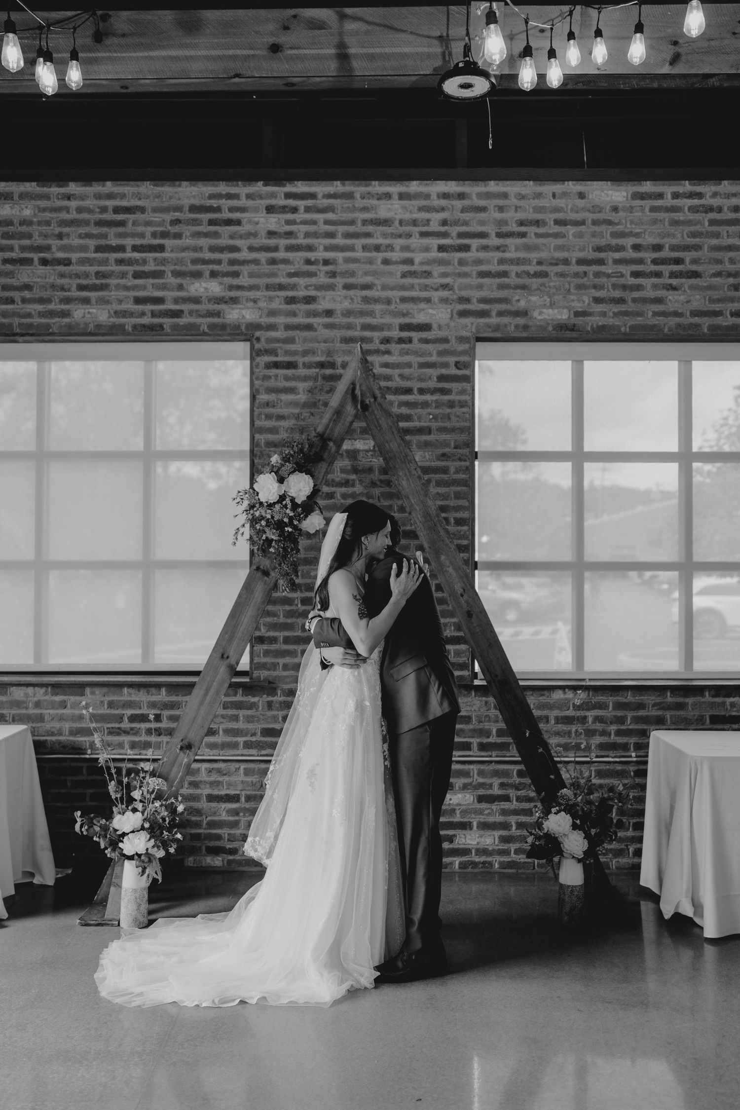 Black and white photo of bride and groom sharing first kiss under triangular wooden arch at indoor wedding ceremony.