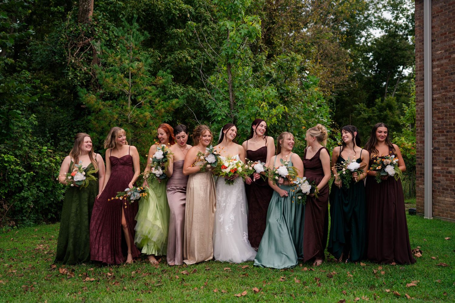 Bride stands with her bridesmaids in mismatched earth-tone dresses holding bouquets outdoors.