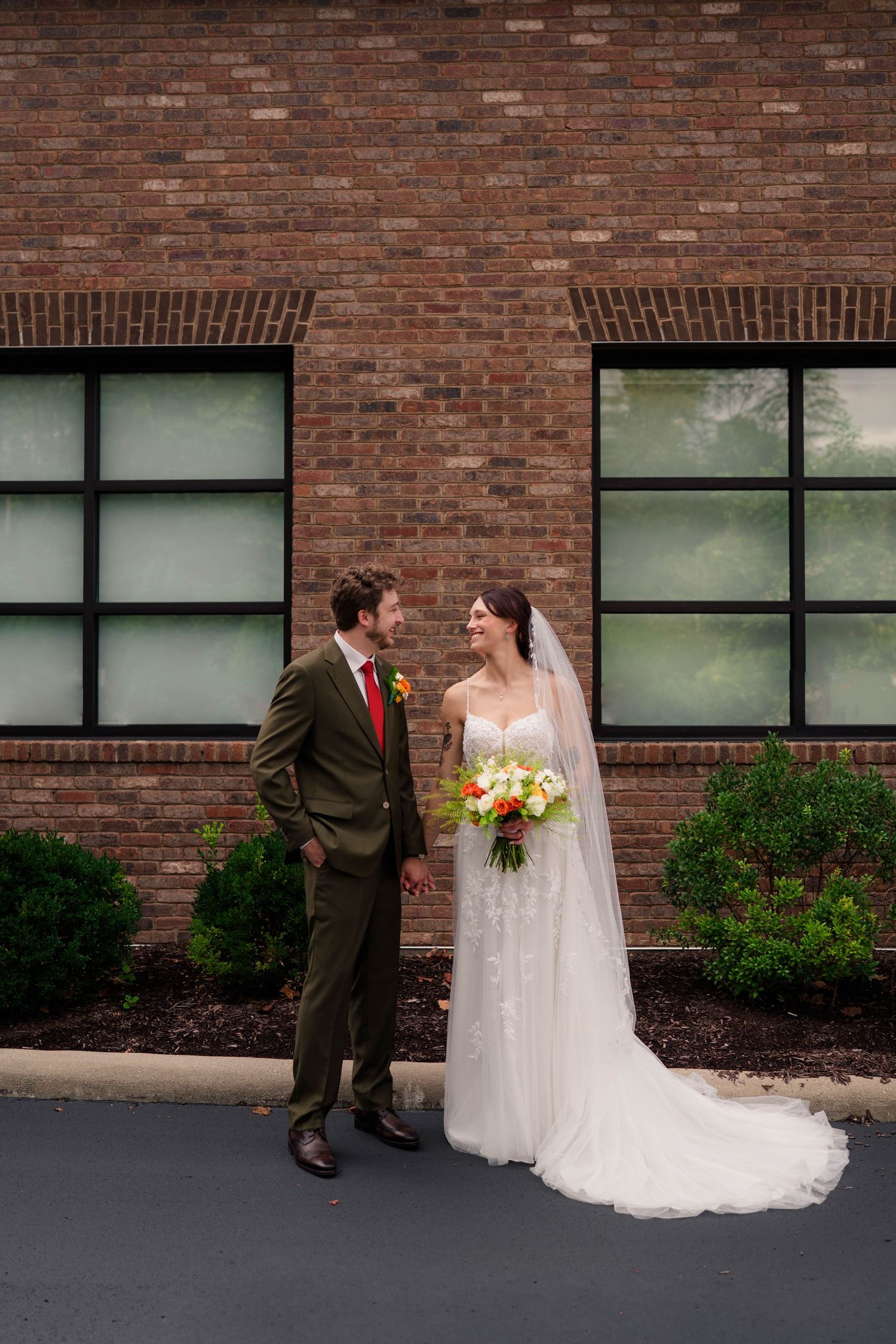 Bride and groom smiling at each other outside brick building, bride holding colorful bouquet.