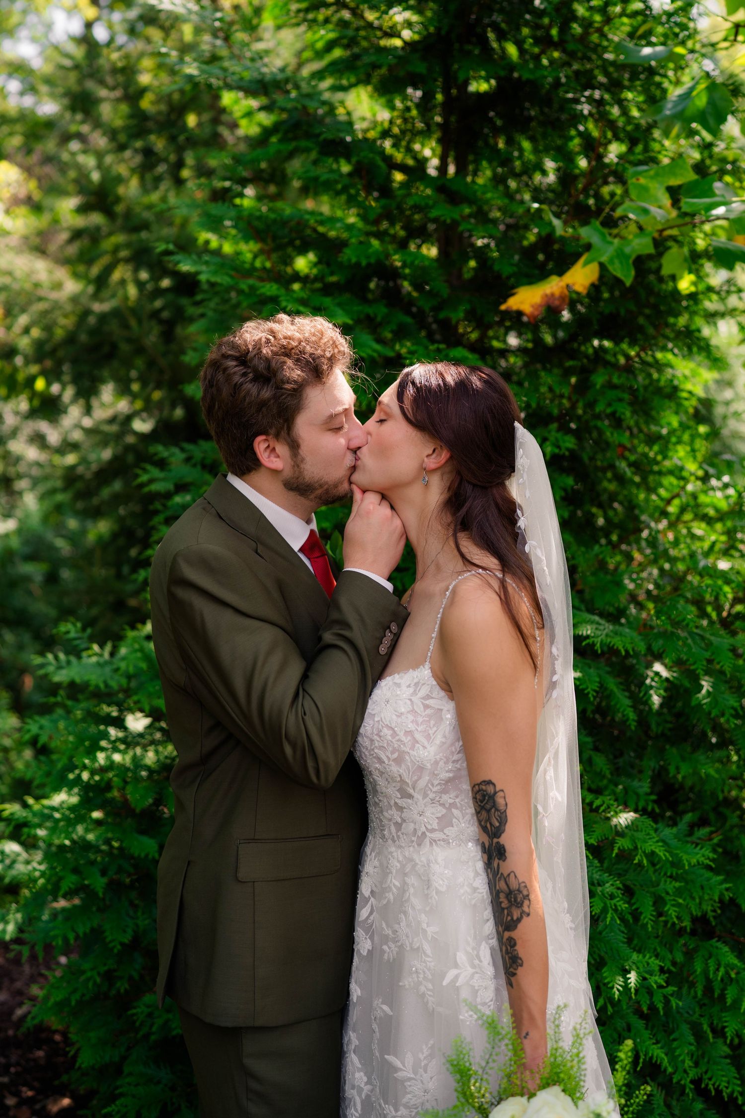 Bride and groom share romantic kiss surrounded by lush green trees in outdoor garden setting.