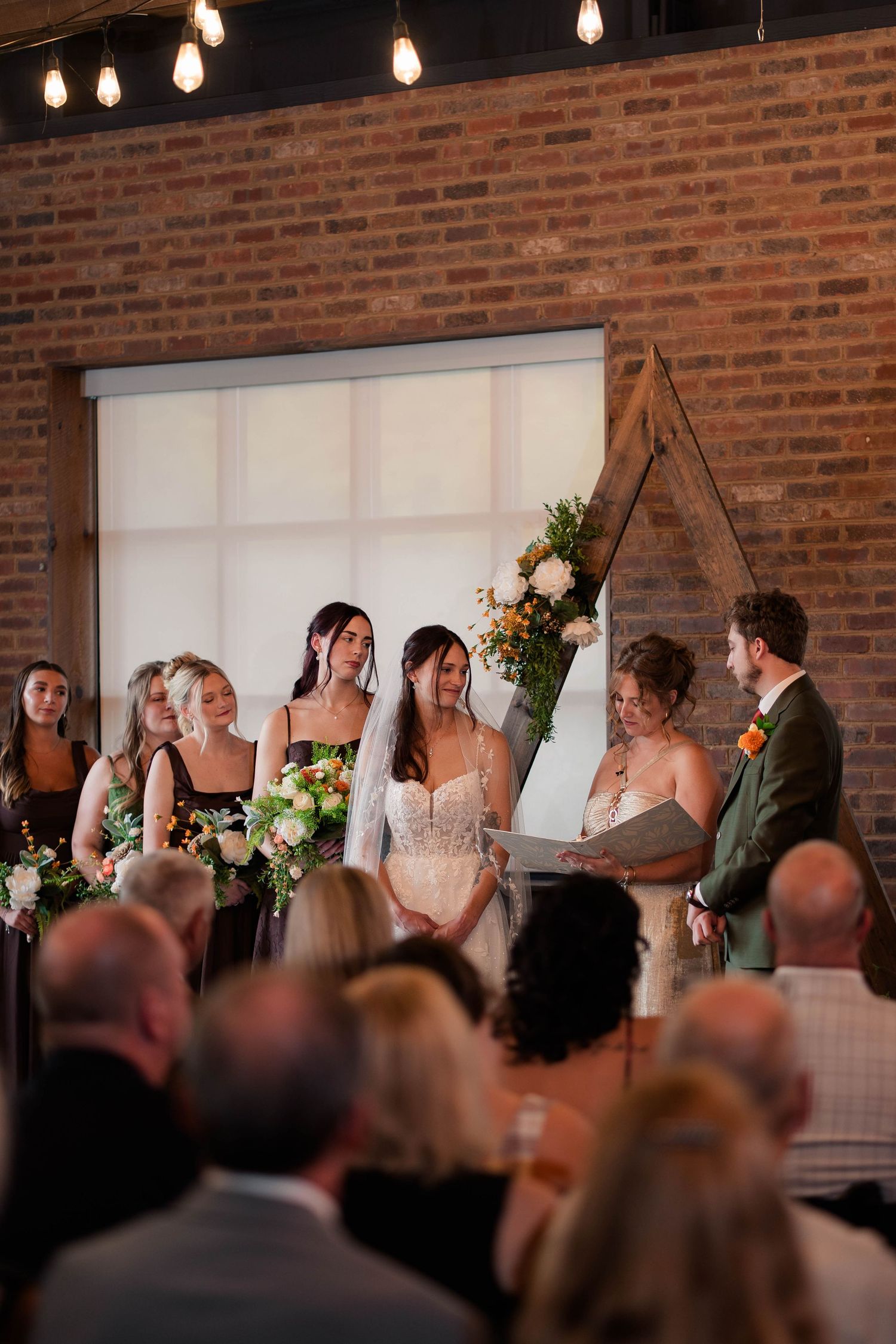Bride in lace gown stands at altar with bridesmaids in black dresses during indoor wedding ceremony.